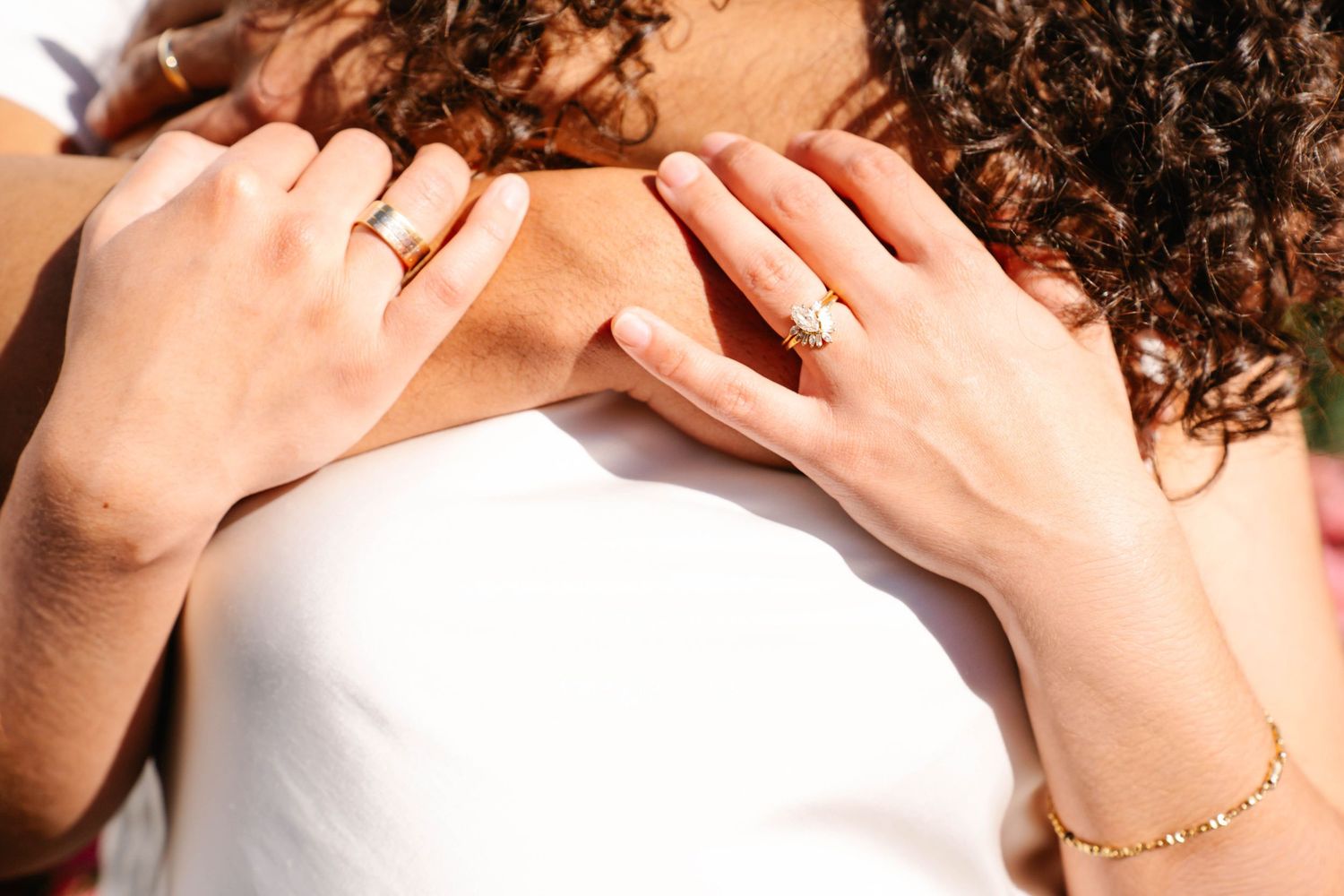 Close-up of two hands with gold rings and bracelet resting on a white wedding dress.