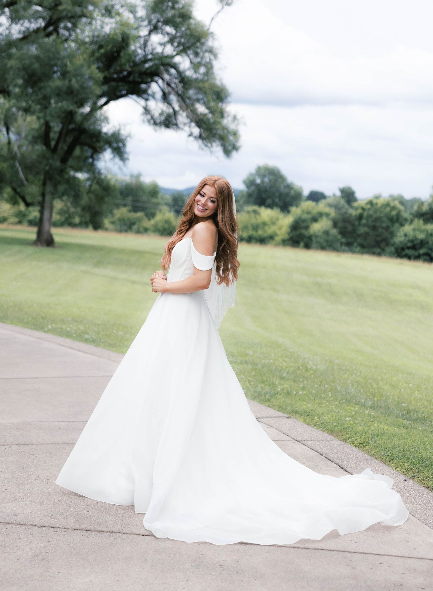 Bride in off-shoulder white gown with flowing train smiles over shoulder on outdoor path with green landscape.