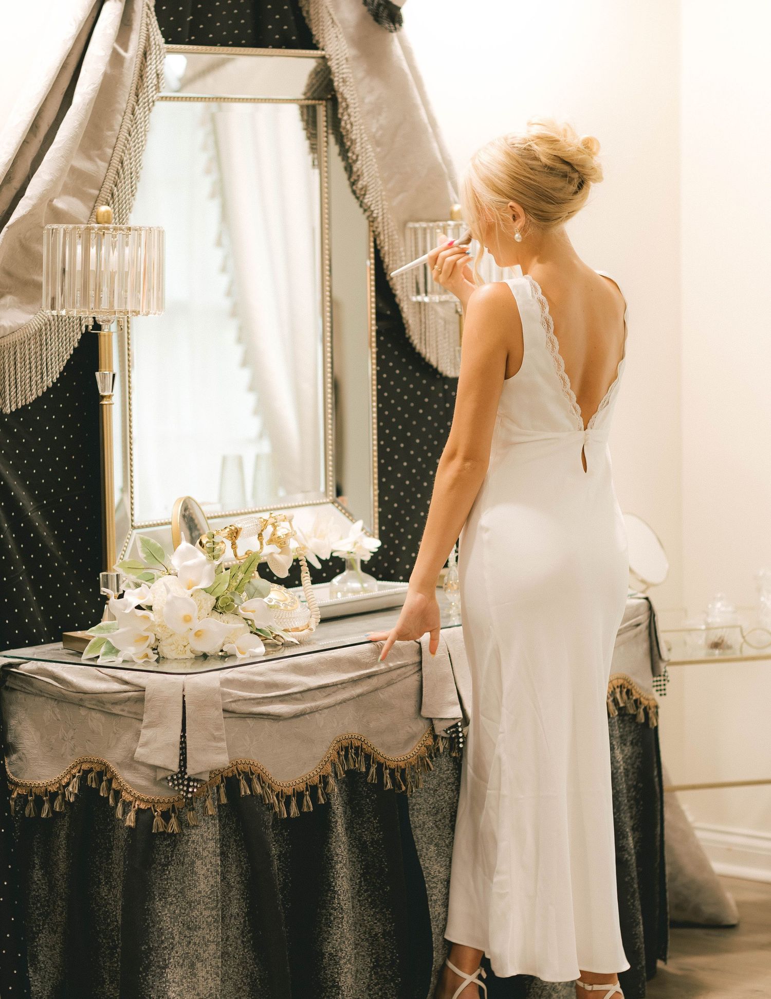 Elegant bride in white lace-back slip dress applying makeup at a vintage vanity mirror with floral arrangement.