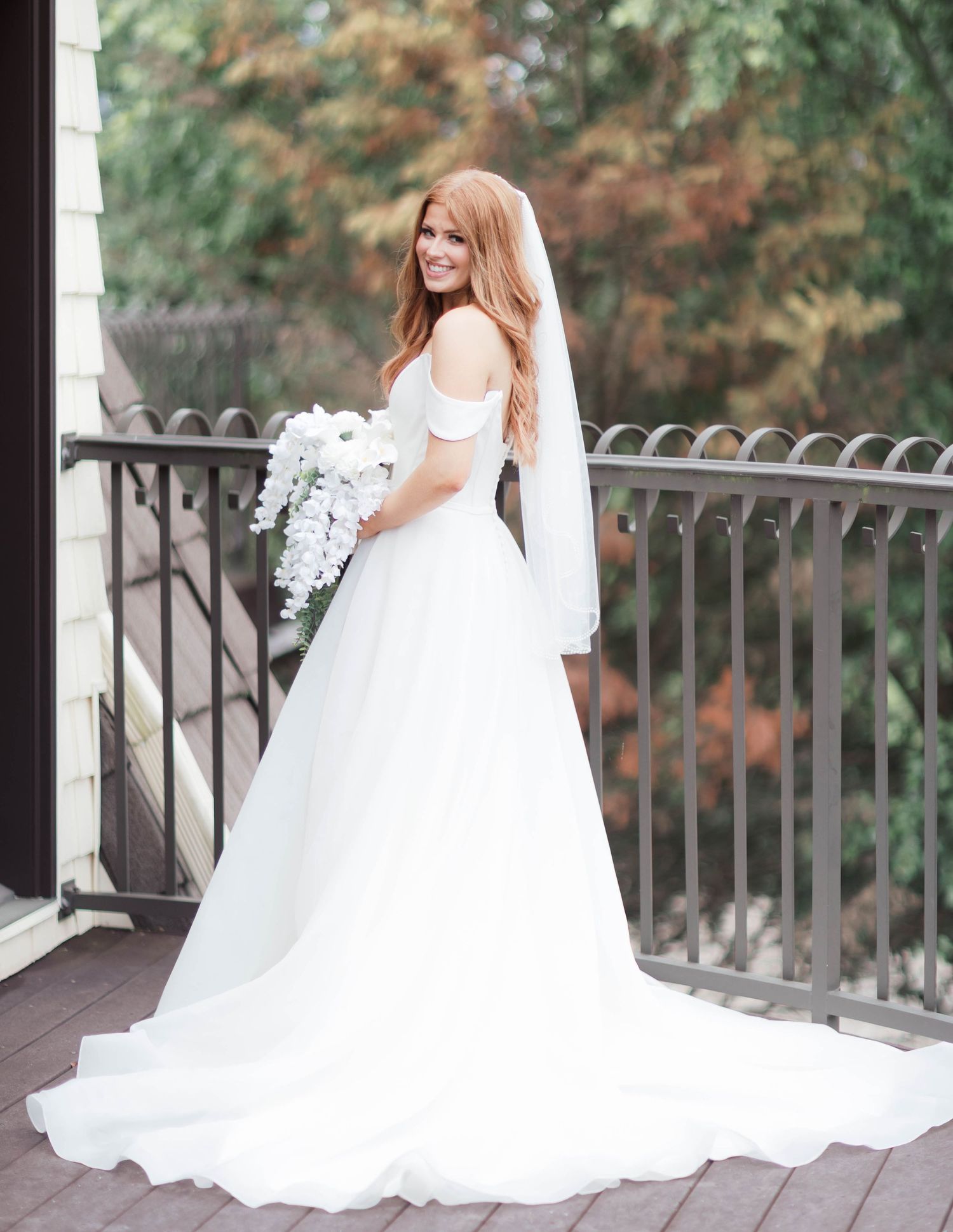 Bride in off-shoulder white ball gown with veil holding white bouquet on outdoor balcony.
