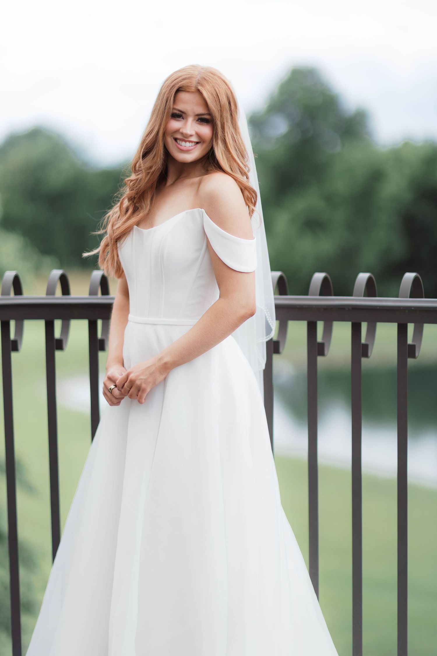 Smiling bride in white off-shoulder wedding gown with veil, posing on outdoor balcony with green trees in background.