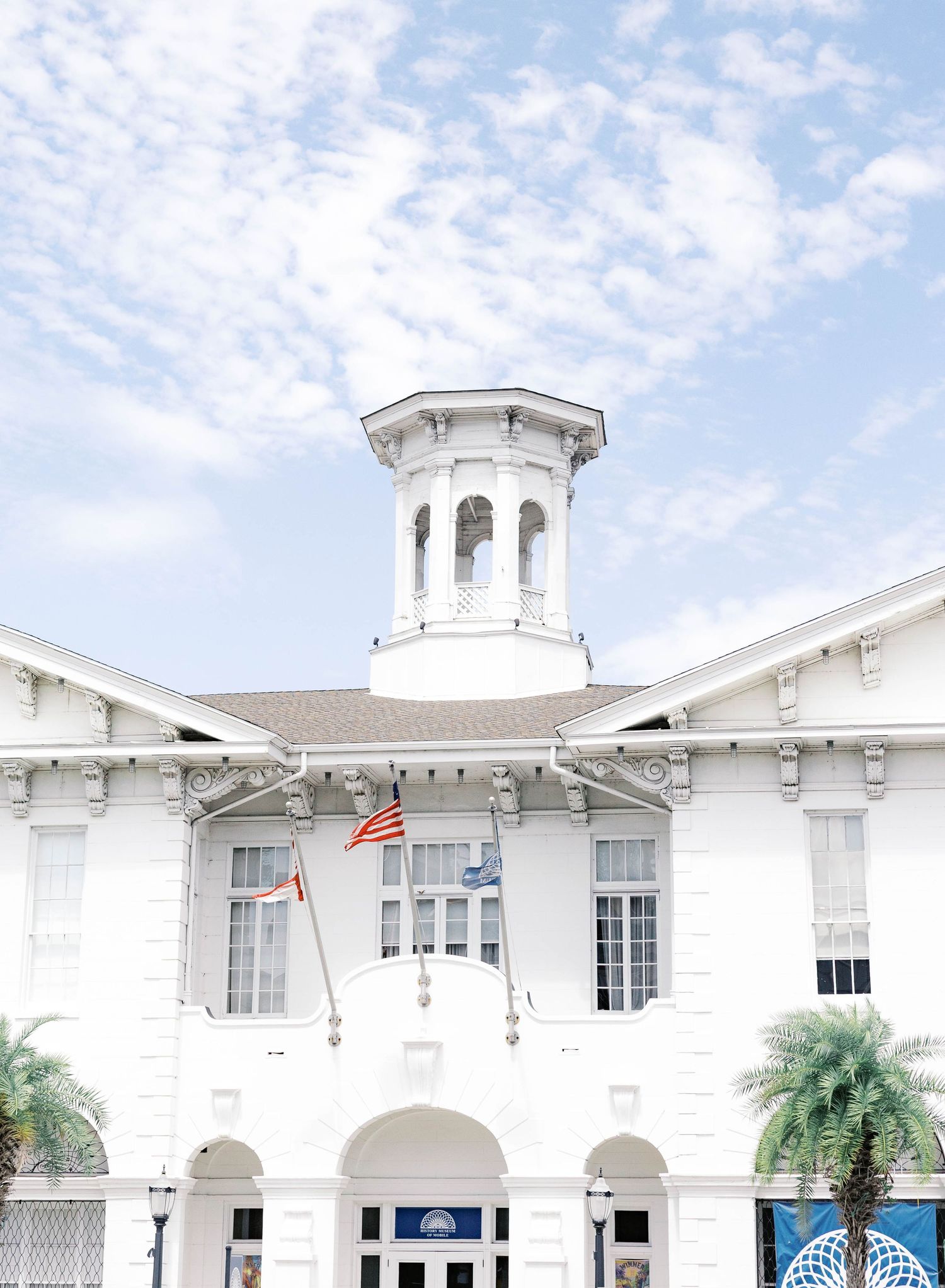 White historic building with ornate cupola tower, American flag, and palm trees under a partly cloudy blue sky.