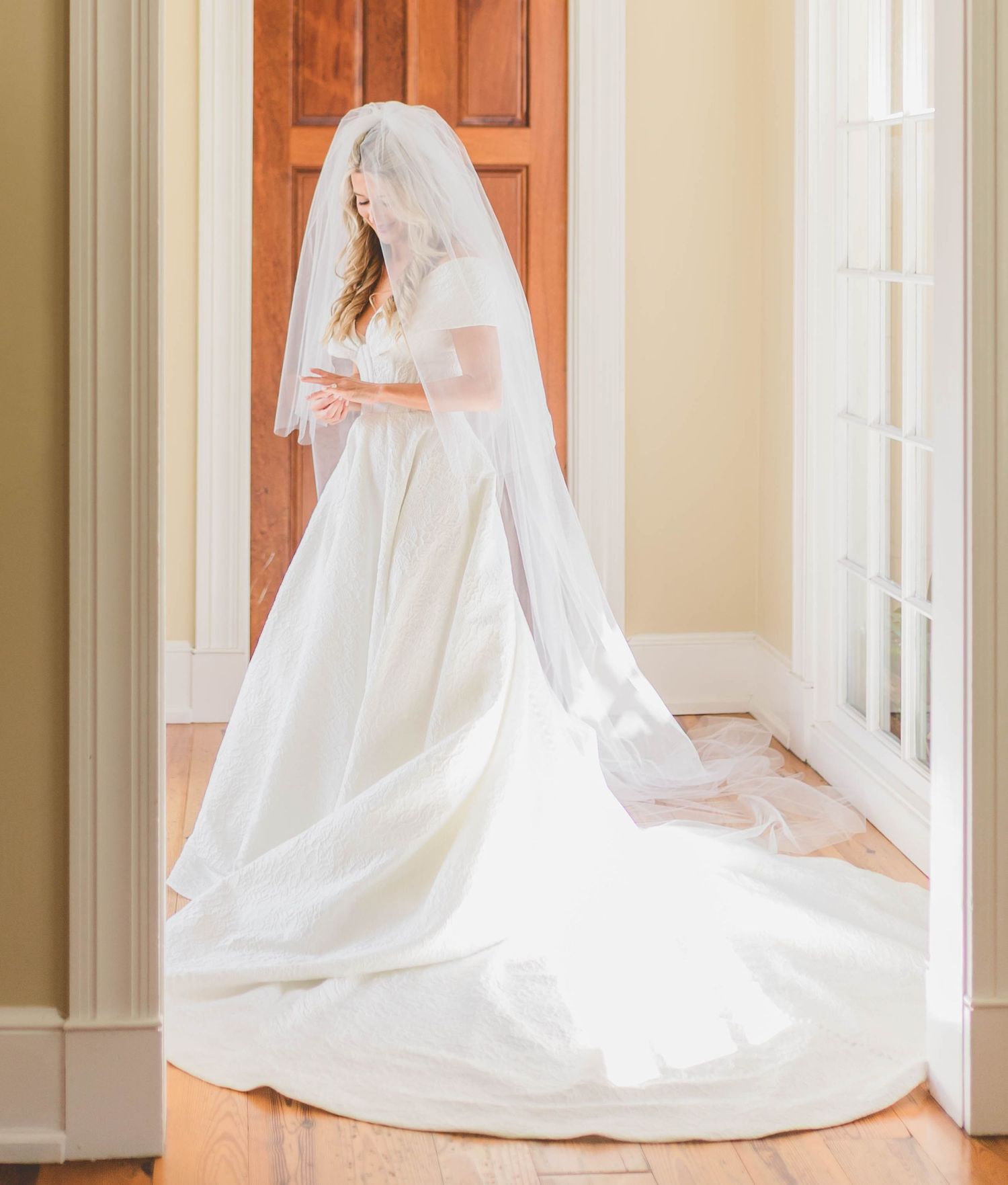 Bride in elegant white wedding gown with long veil and cathedral train standing in a doorway.