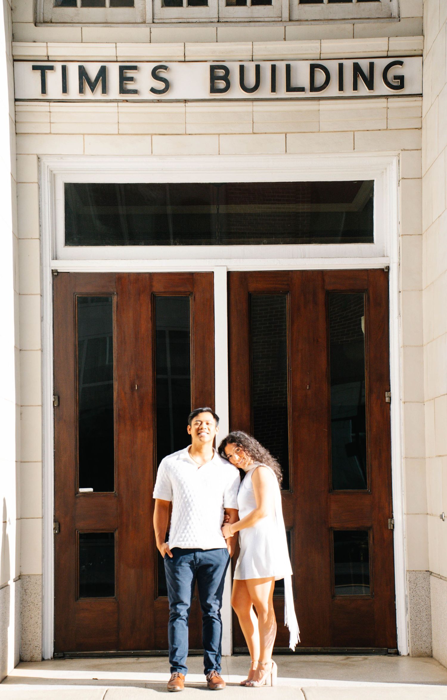 Couple posing in front of the Times Building entrance with wooden doors, man in white shirt and woman in white dress.
