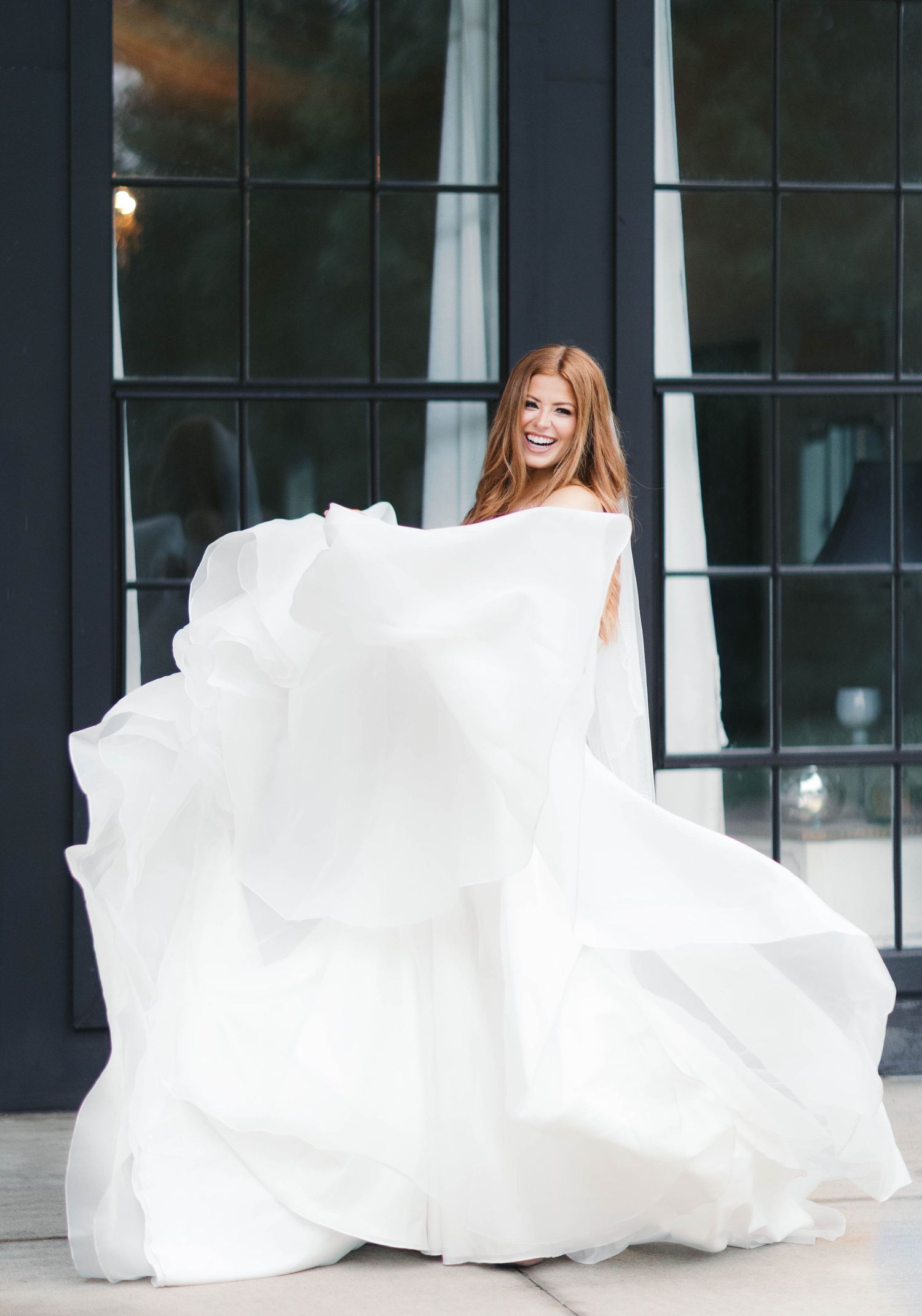 Joyful redhead bride twirling her voluminous white ball gown outside a modern venue.