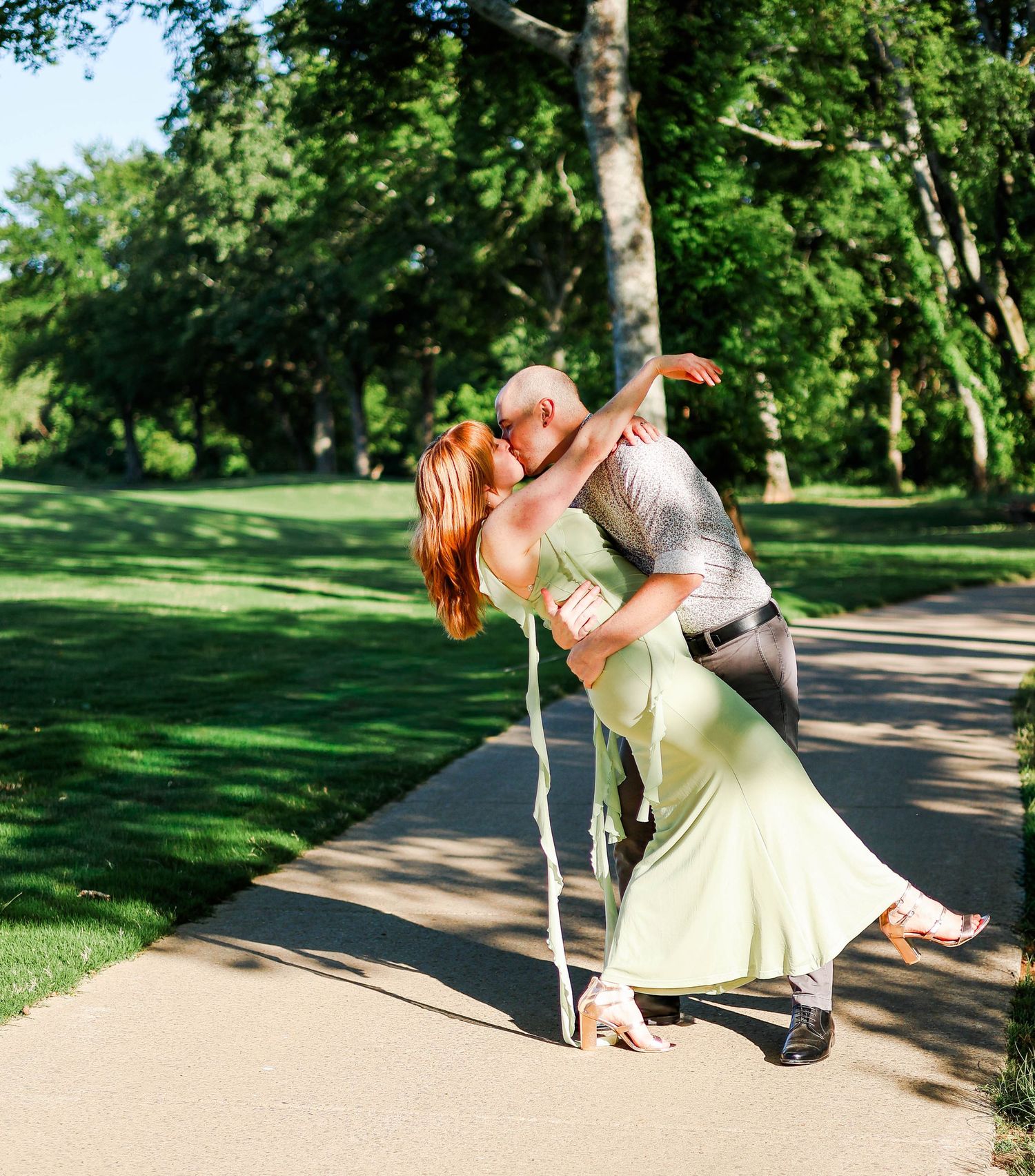 Couple sharing a romantic dip kiss on a tree-lined path, woman in green dress, man in dress shirt and slacks.