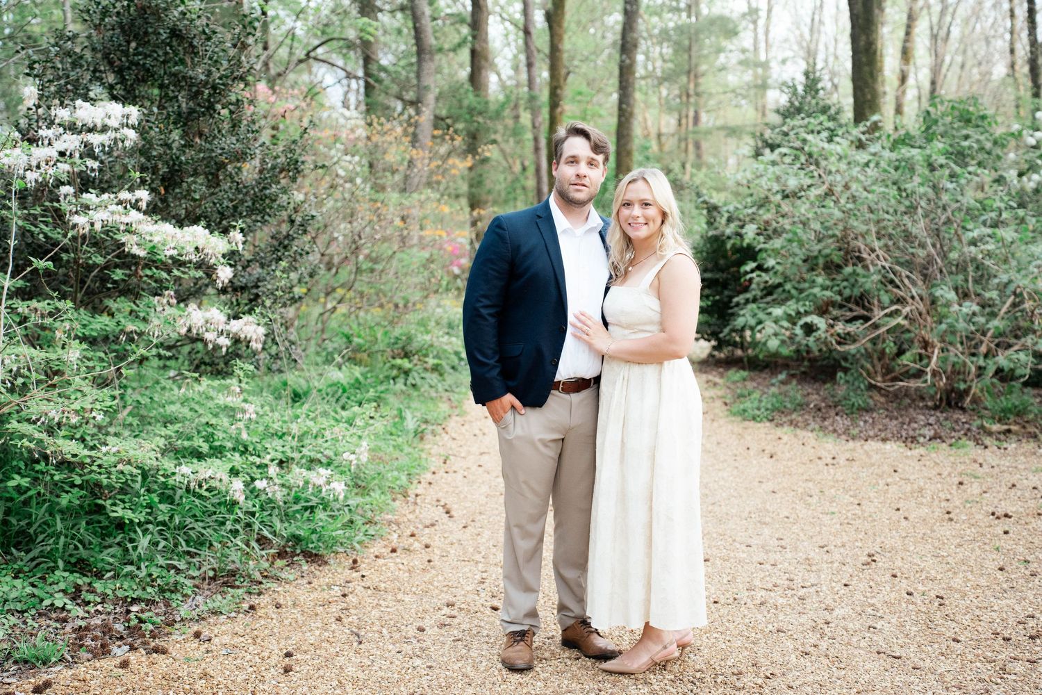 Couple posing together on a garden path surrounded by blooming trees and lush greenery.