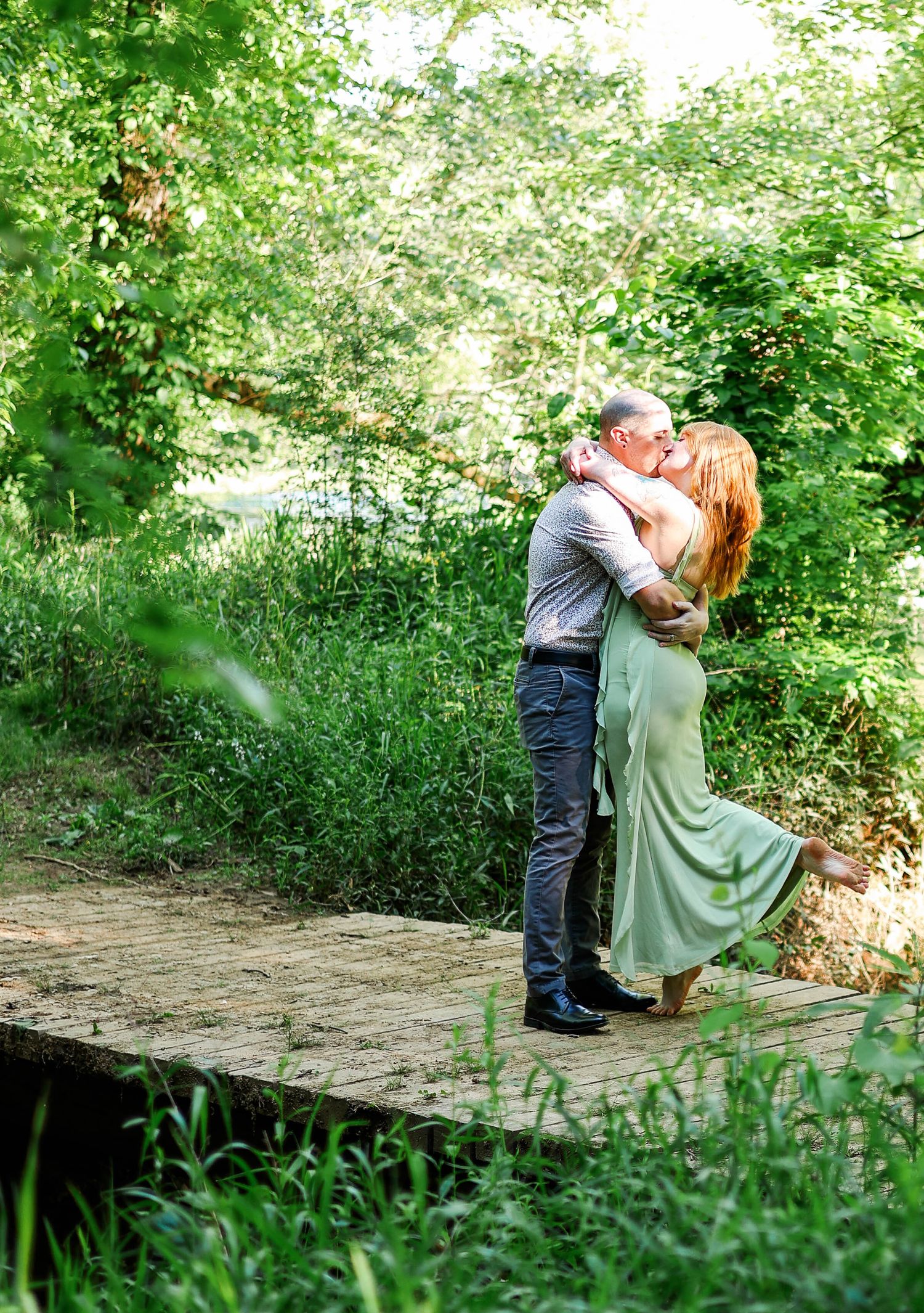 Couple sharing a romantic kiss on a wooden bridge surrounded by lush green trees and foliage outdoors.
