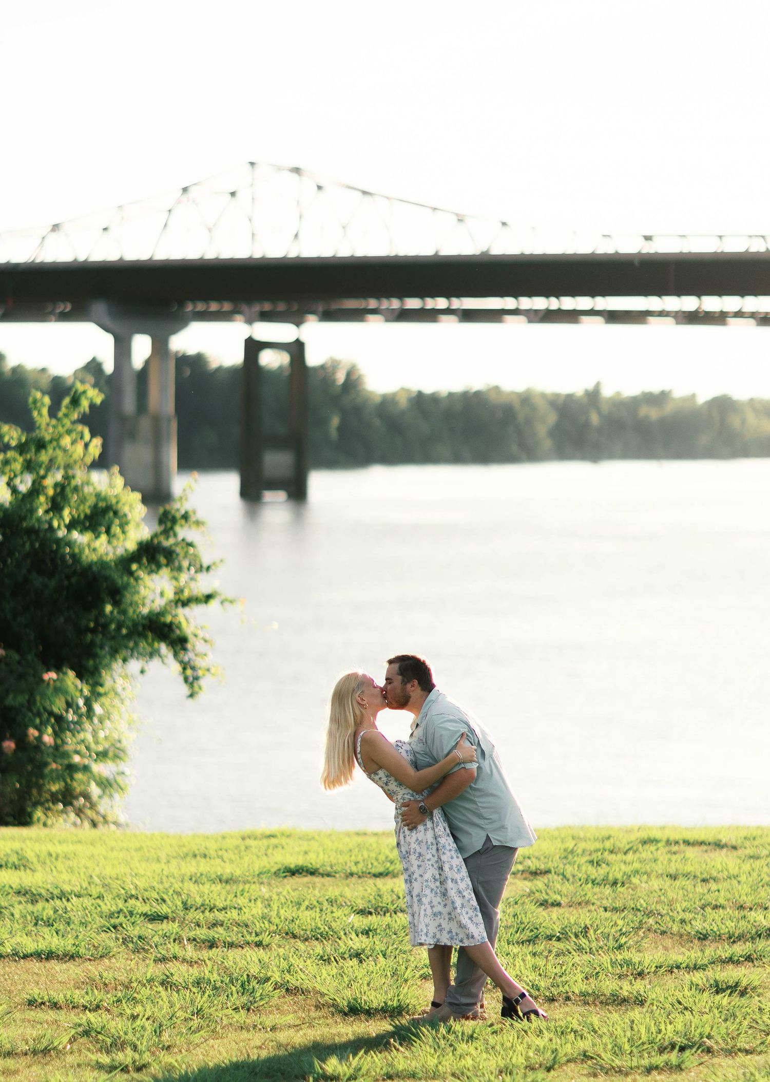 Couple sharing a romantic kiss on green grass with a large river bridge and water in the background at sunset.