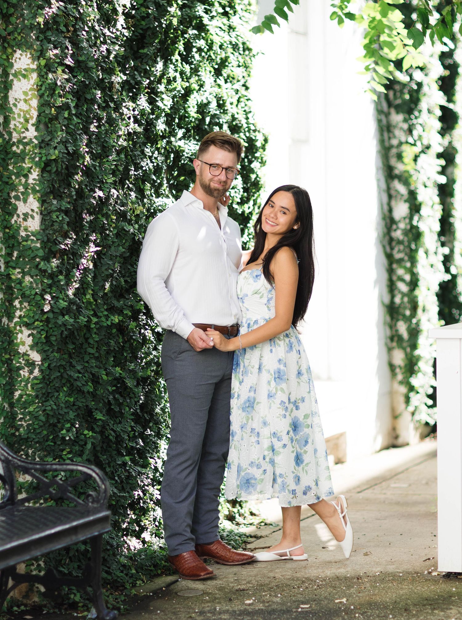 Couple posing together outdoors near ivy-covered wall, man in white shirt and woman in floral dress, elegant engagement photo.