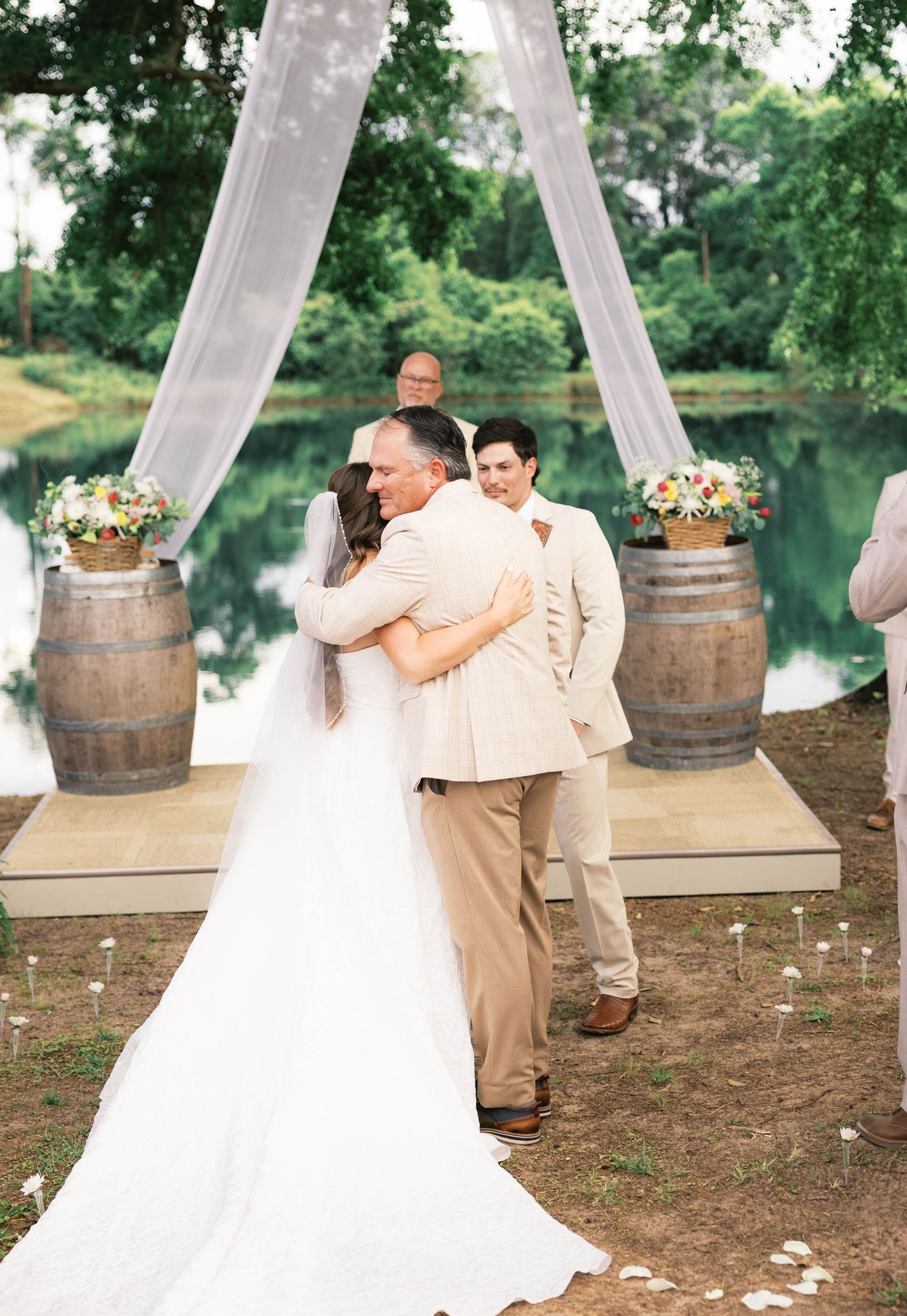 Bride and groom share first kiss at outdoor wedding ceremony with rustic wine barrel decor and sheer white draping by a pond.