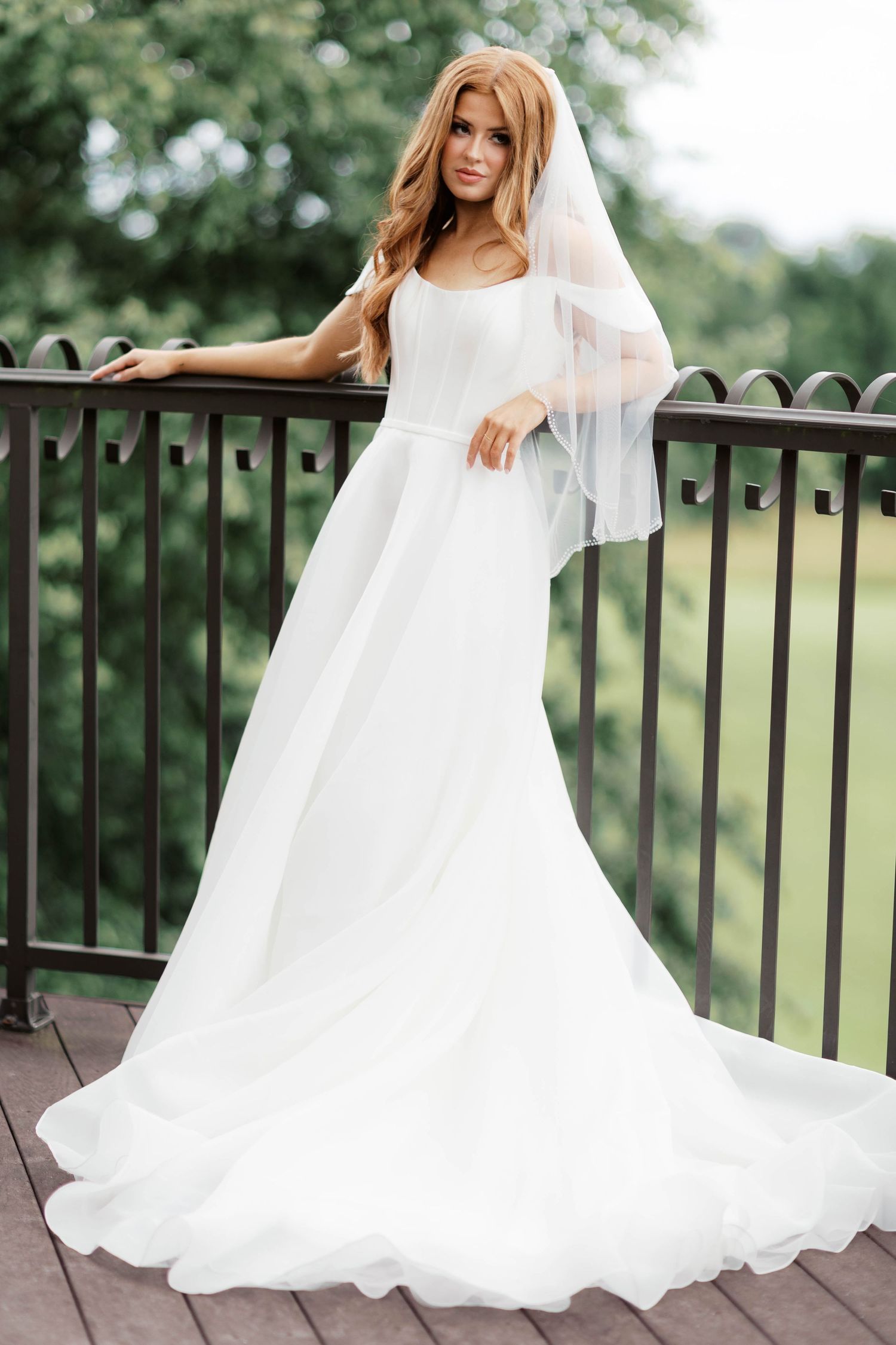 Elegant bride in white A-line gown with veil poses on wooden balcony surrounded by lush greenery.
