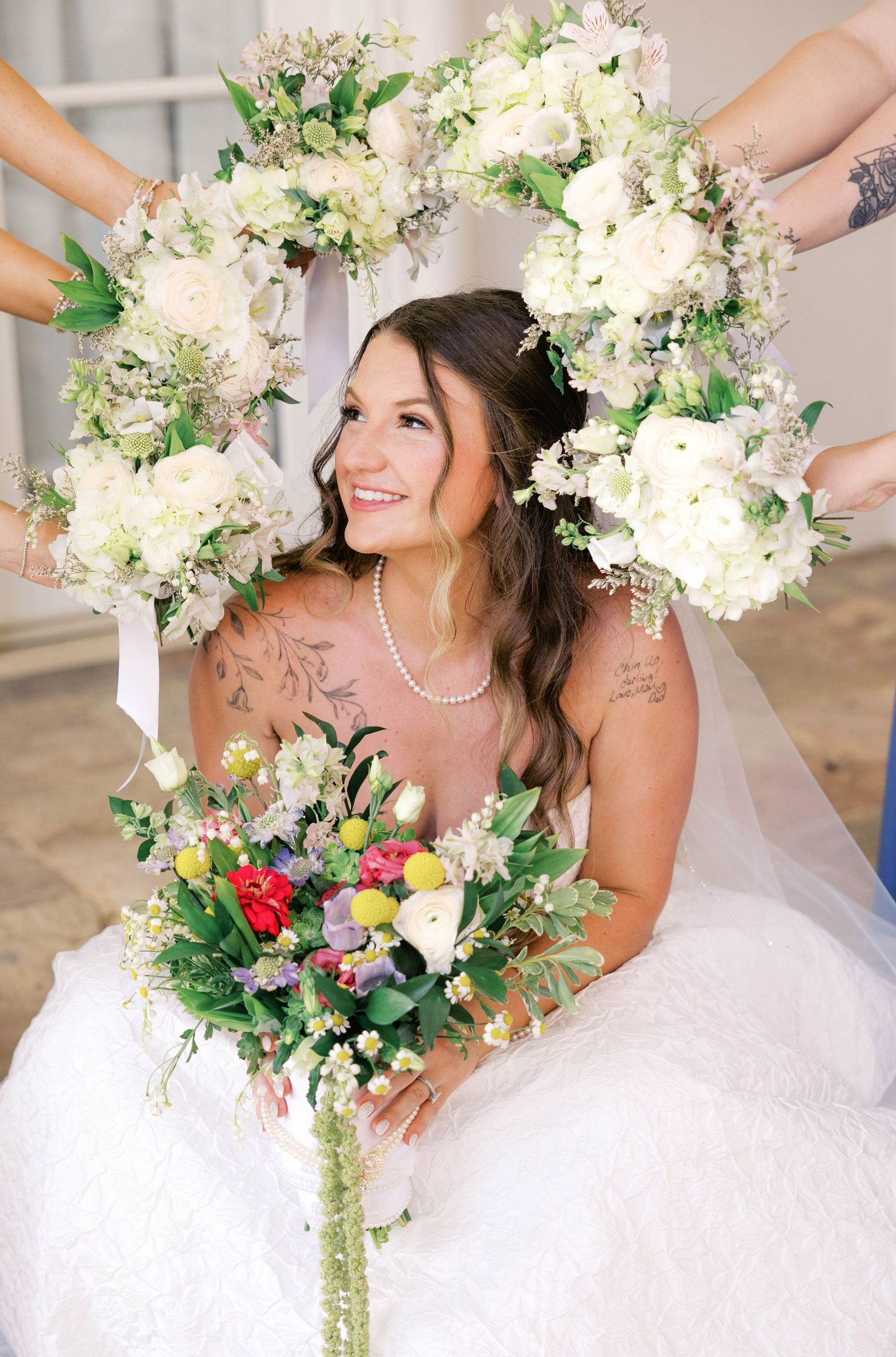 Smiling bride in white gown holds colorful bouquet, framed by floral wreath of white roses held by bridesmaids.