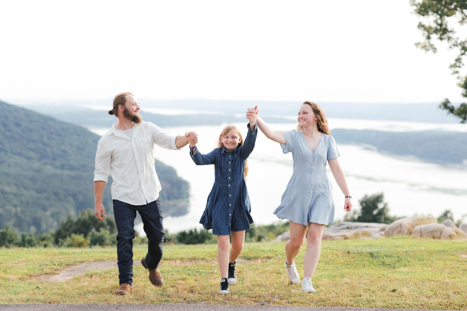 A happy family of three holds hands and walks outdoors with mountain and river views in the background.
