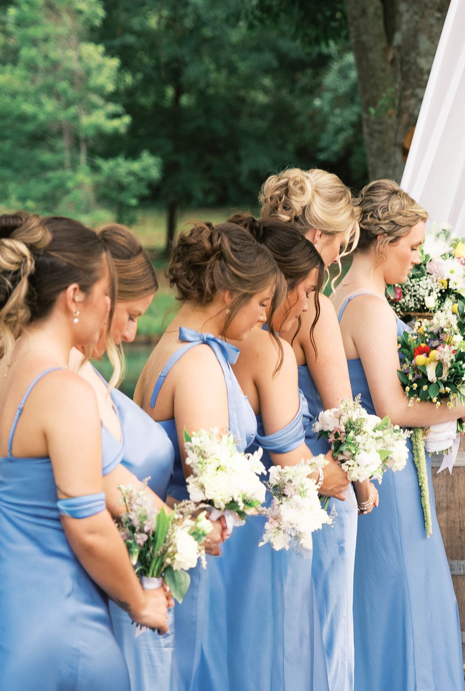Bridesmaids in blue dresses holding white floral bouquets standing in a row at an outdoor wedding ceremony.