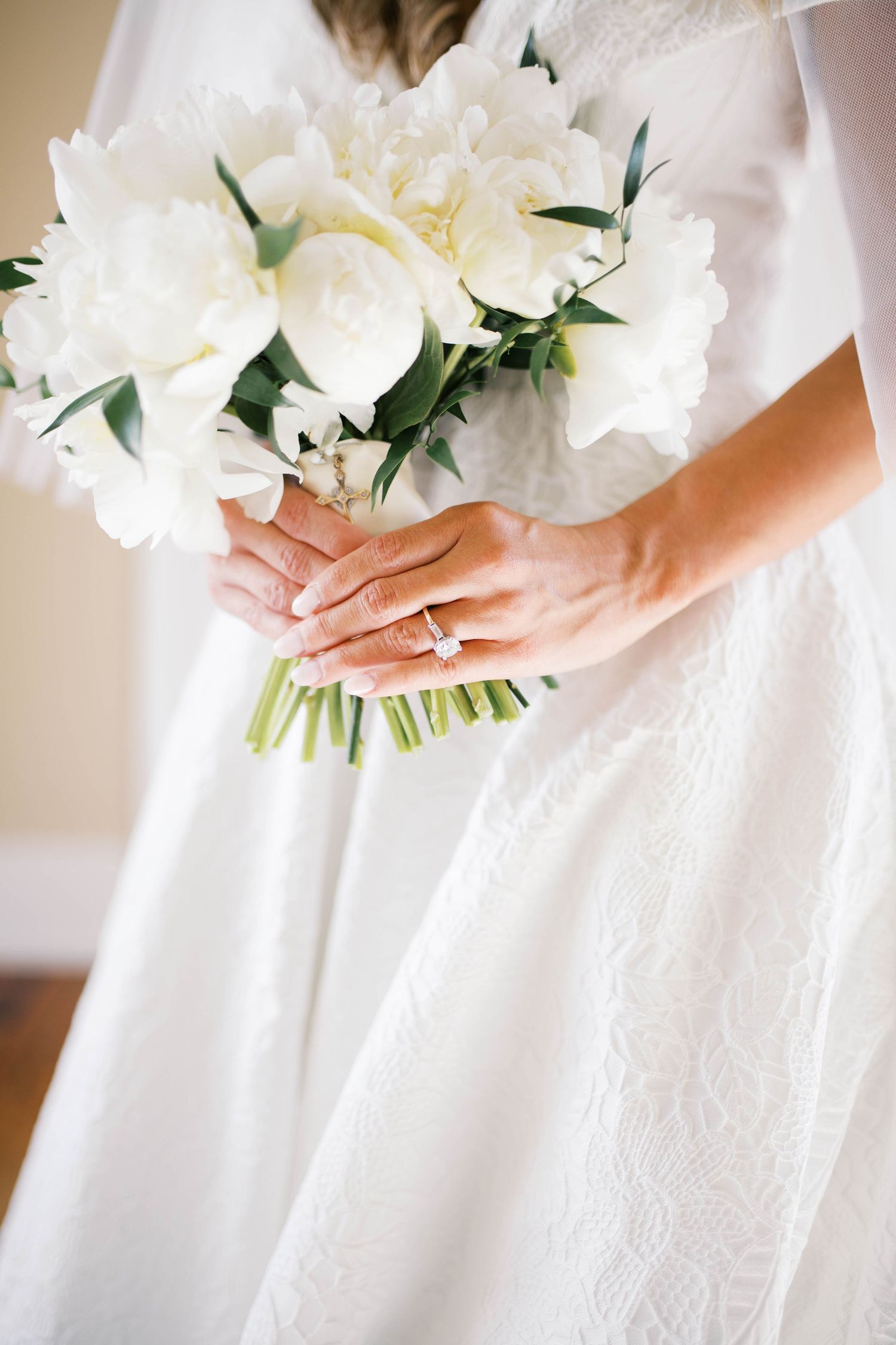 Bride in white lace gown holding elegant bouquet of white peonies, showcasing diamond engagement ring.