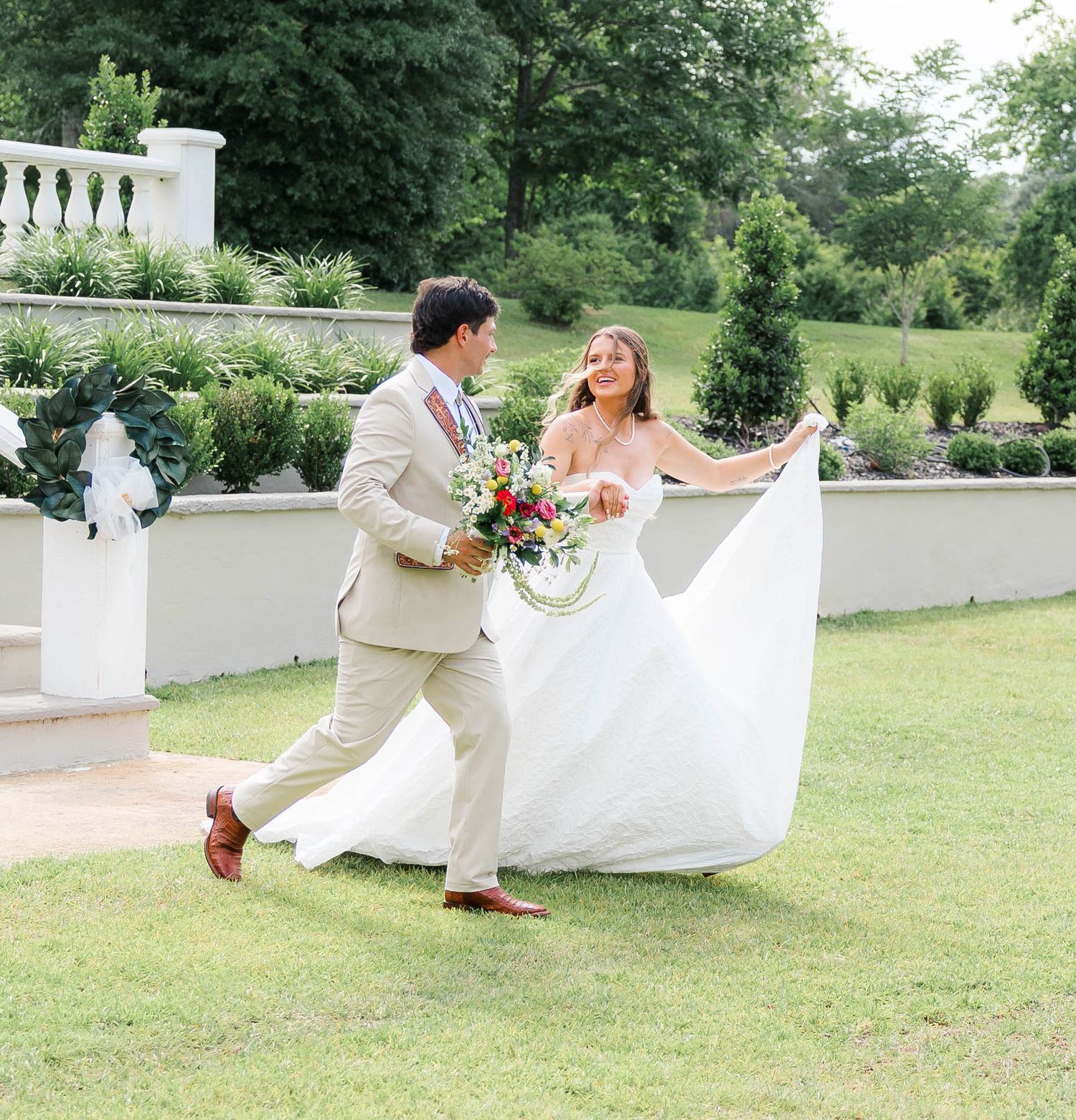 A bride in a flowing white gown and groom in a tan suit walk joyfully across green grass at an outdoor wedding venue.