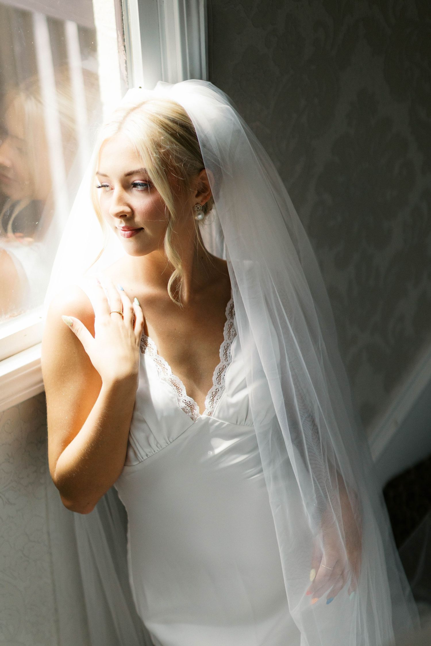 Blonde bride in white lace wedding dress and veil gazing out a sunlit window.