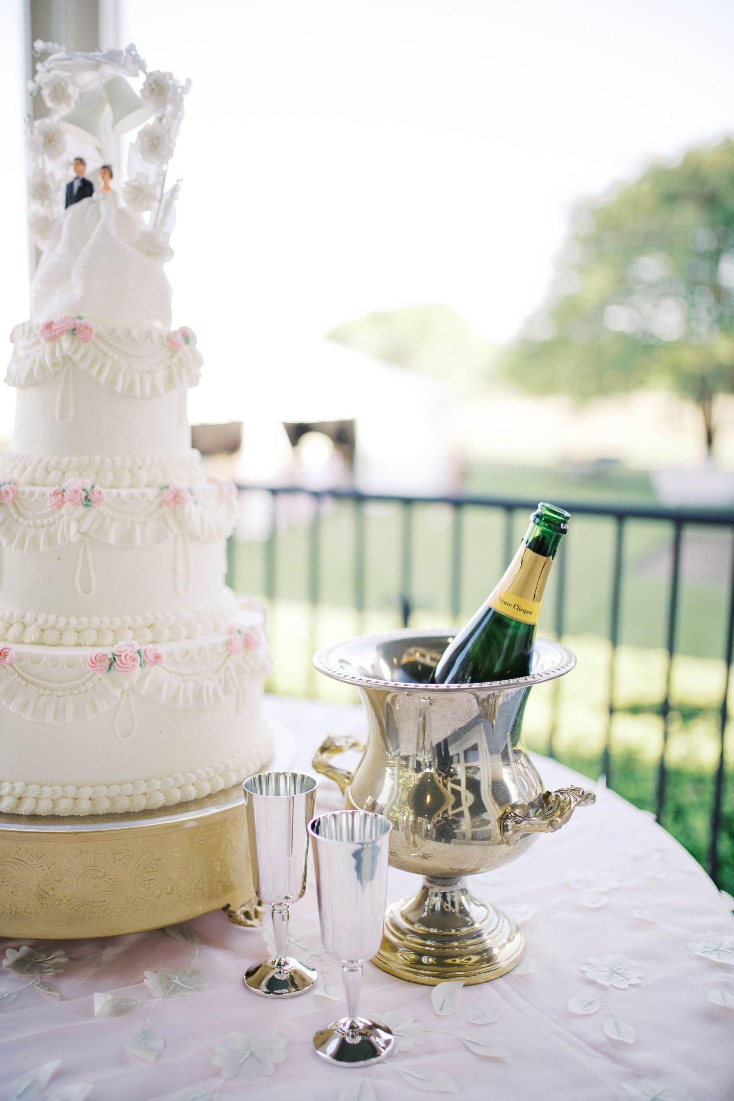 Elegant white tiered wedding cake with bride and groom topper beside champagne bottle in silver ice bucket with flutes.