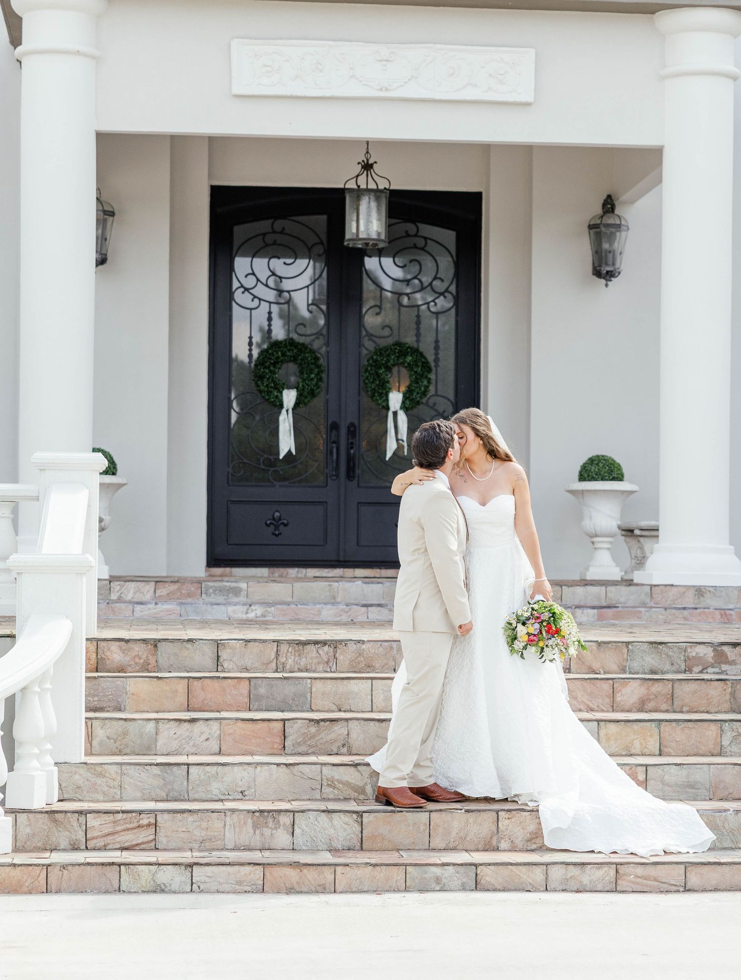 Bride and groom share a kiss on stone steps before elegant white mansion with black double doors and wreaths.
