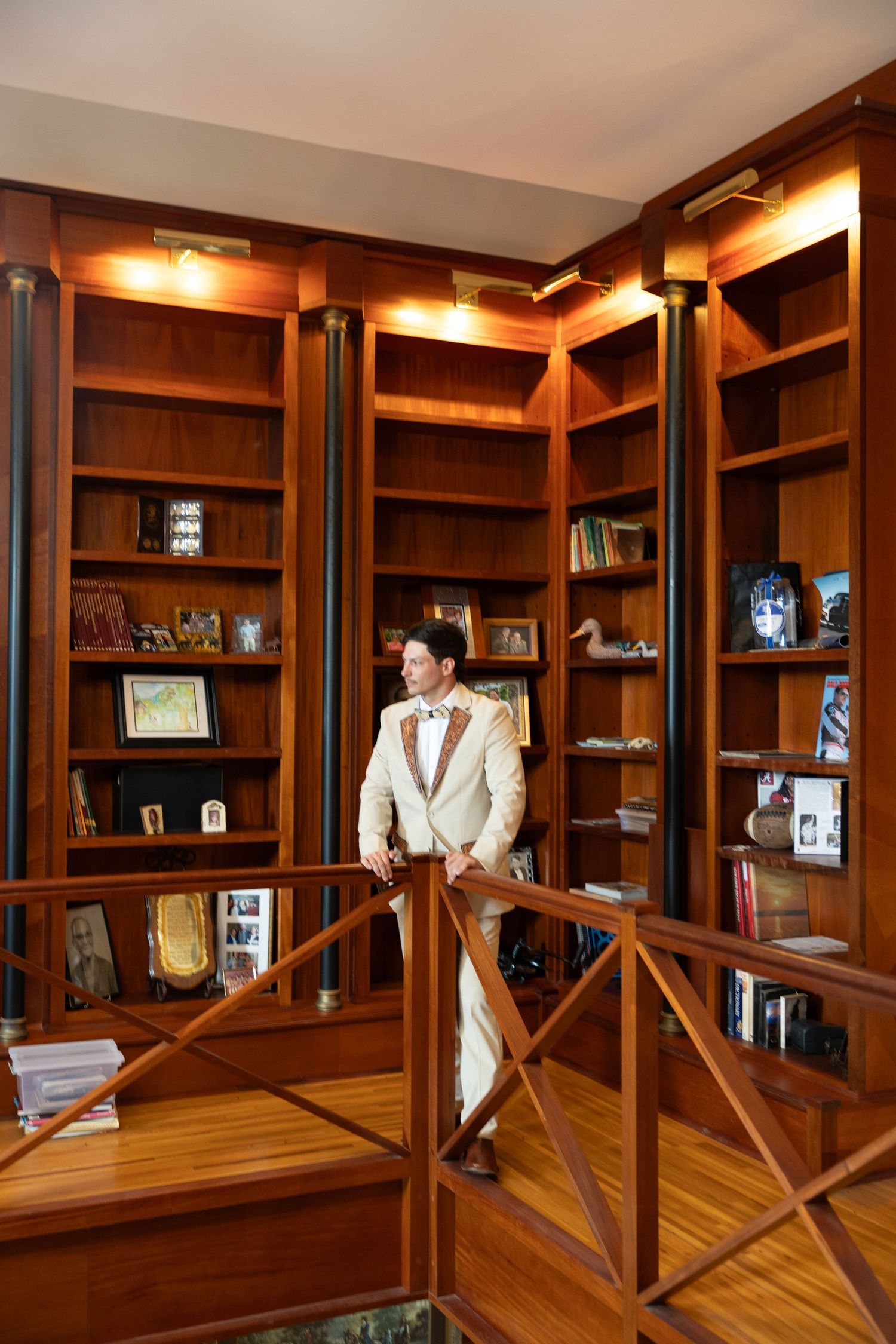 Elegant man in cream suit standing on wooden staircase in front of lit mahogany bookshelves.