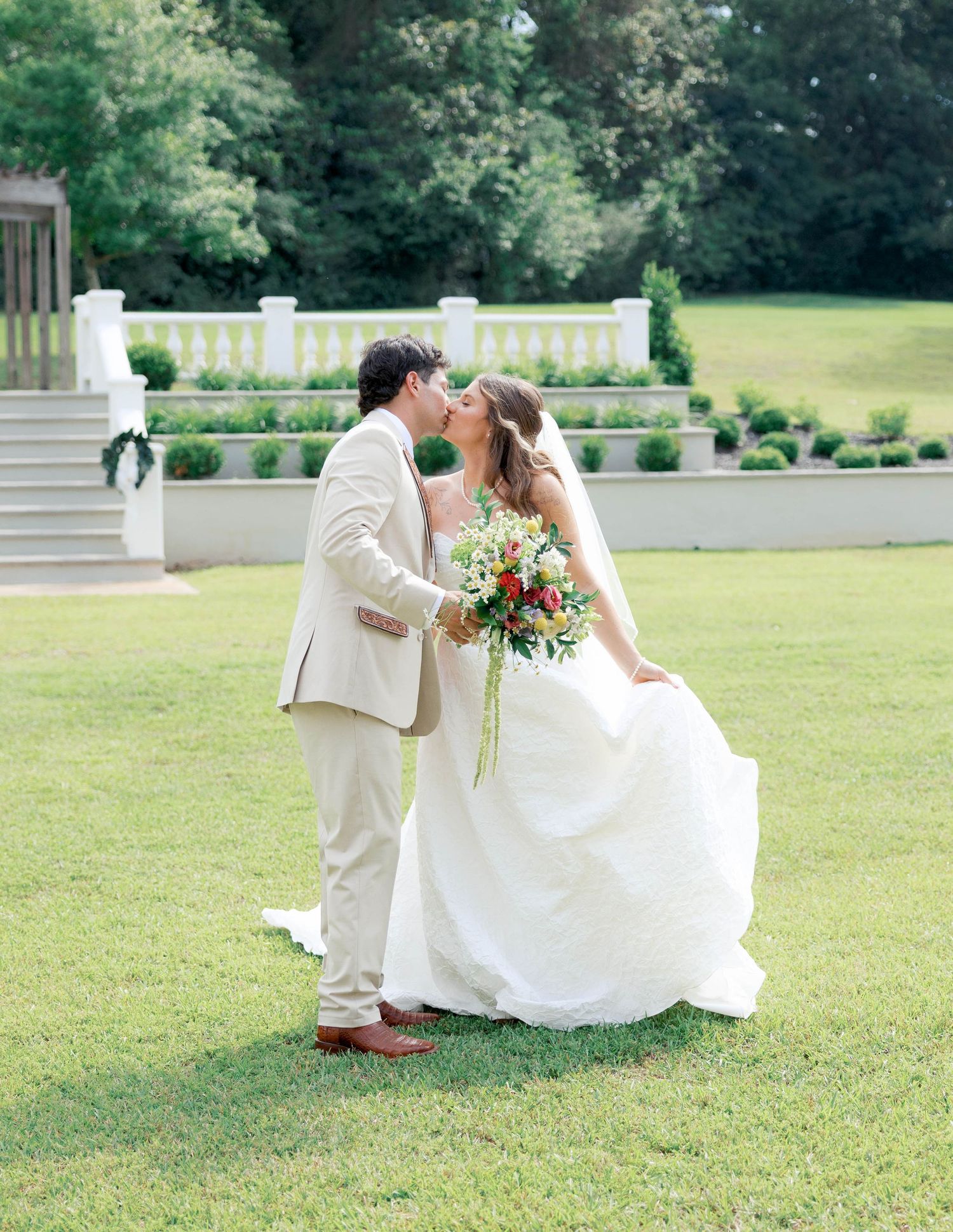 Bride and groom share a kiss on green lawn, she holds colorful bouquet in white gown, he wears tan suit, garden venue.