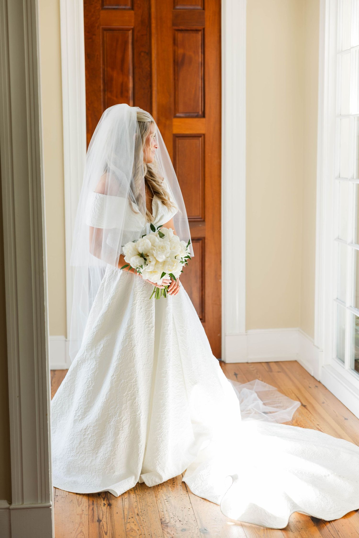 Bride in white lace ball gown with veil and white bouquet standing by wooden doors on hardwood floors.