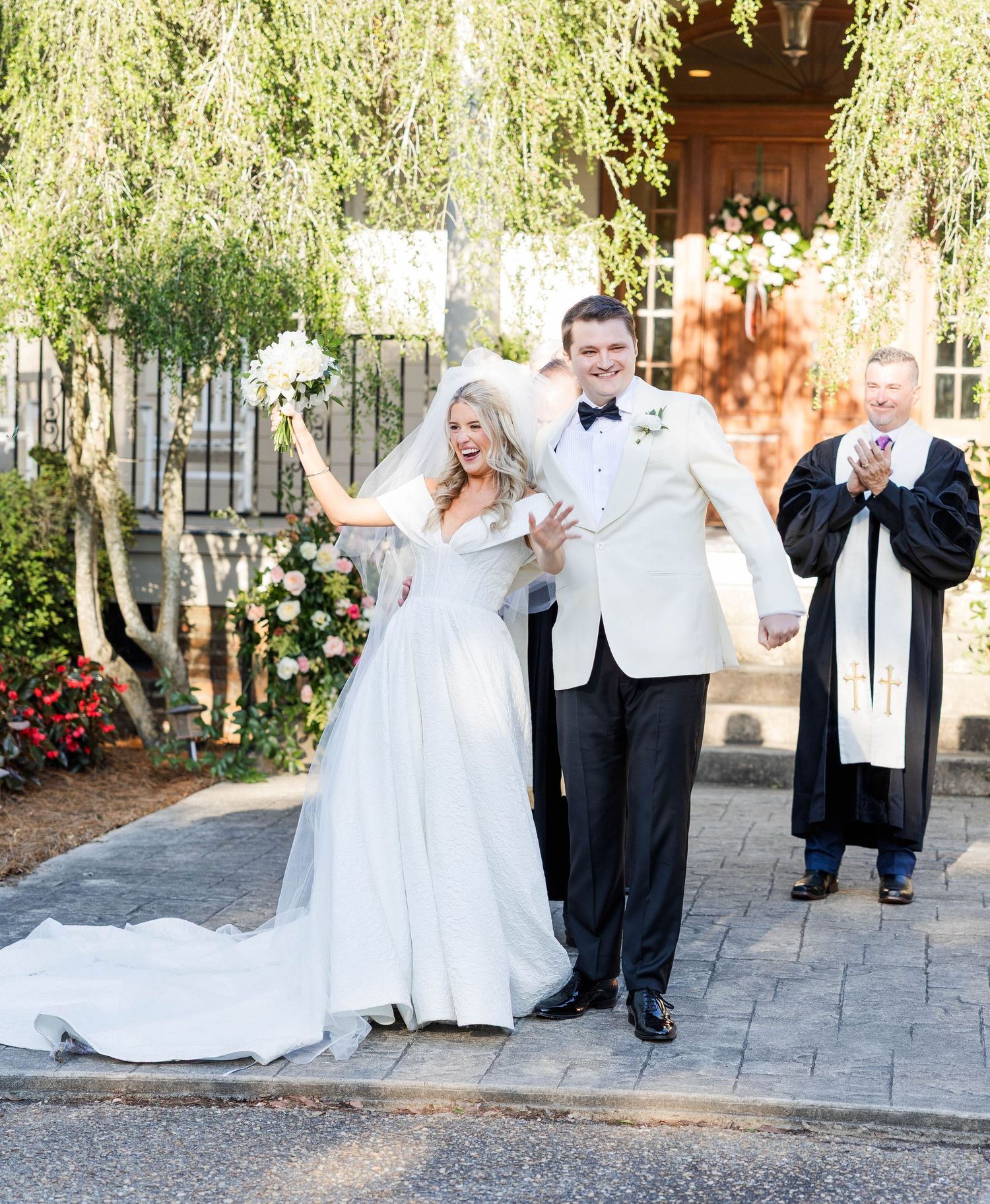 Joyful bride and groom exit wedding ceremony under willow tree, officiant applauding behind them.