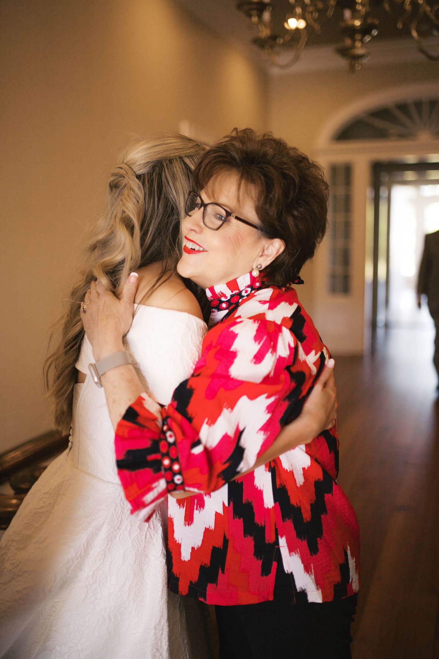 A bride in a white dress shares a warm hug with an older woman in a red patterned jacket inside an elegant home.