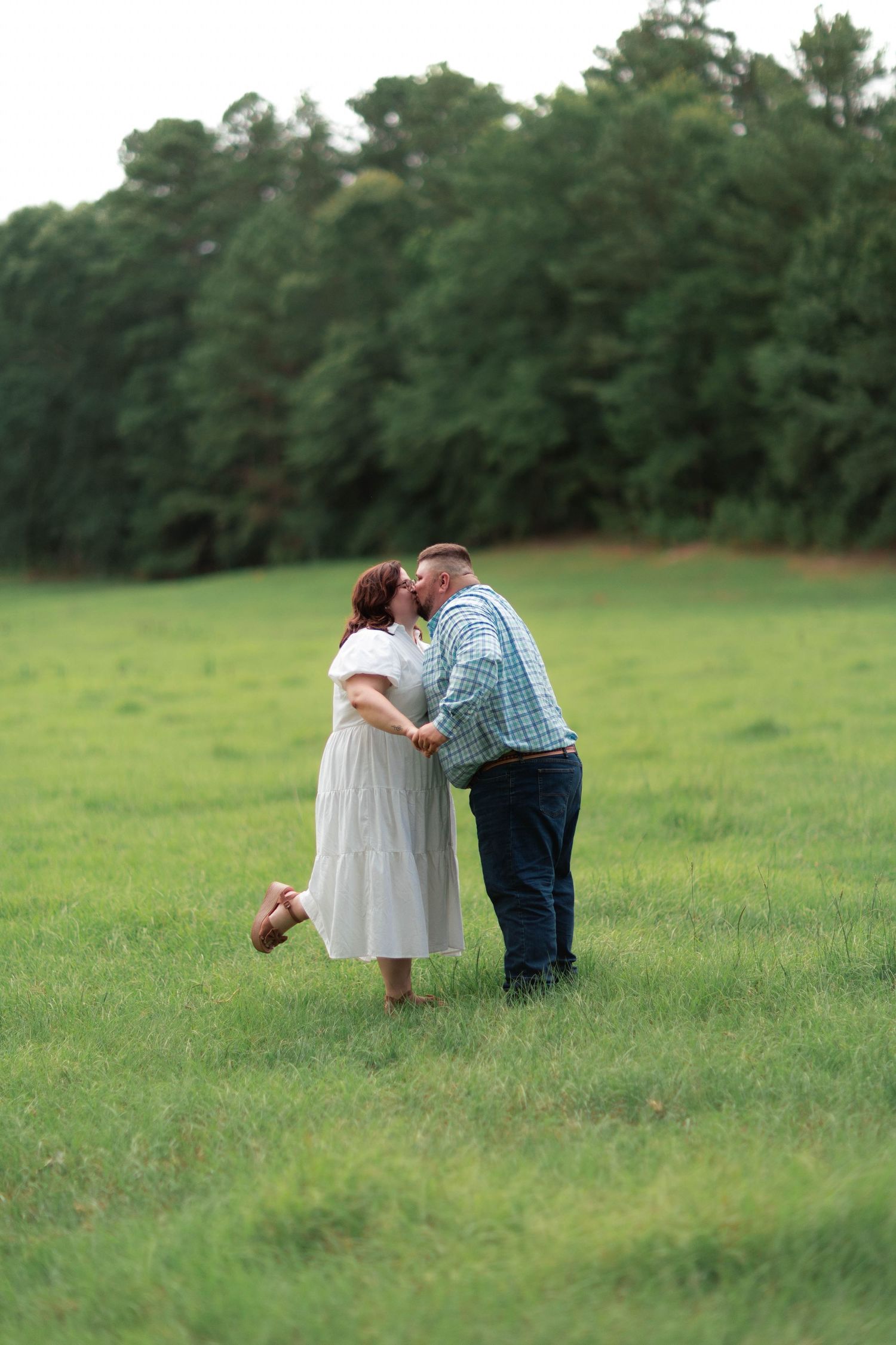 A couple shares a romantic kiss in a lush green field with trees in the background.