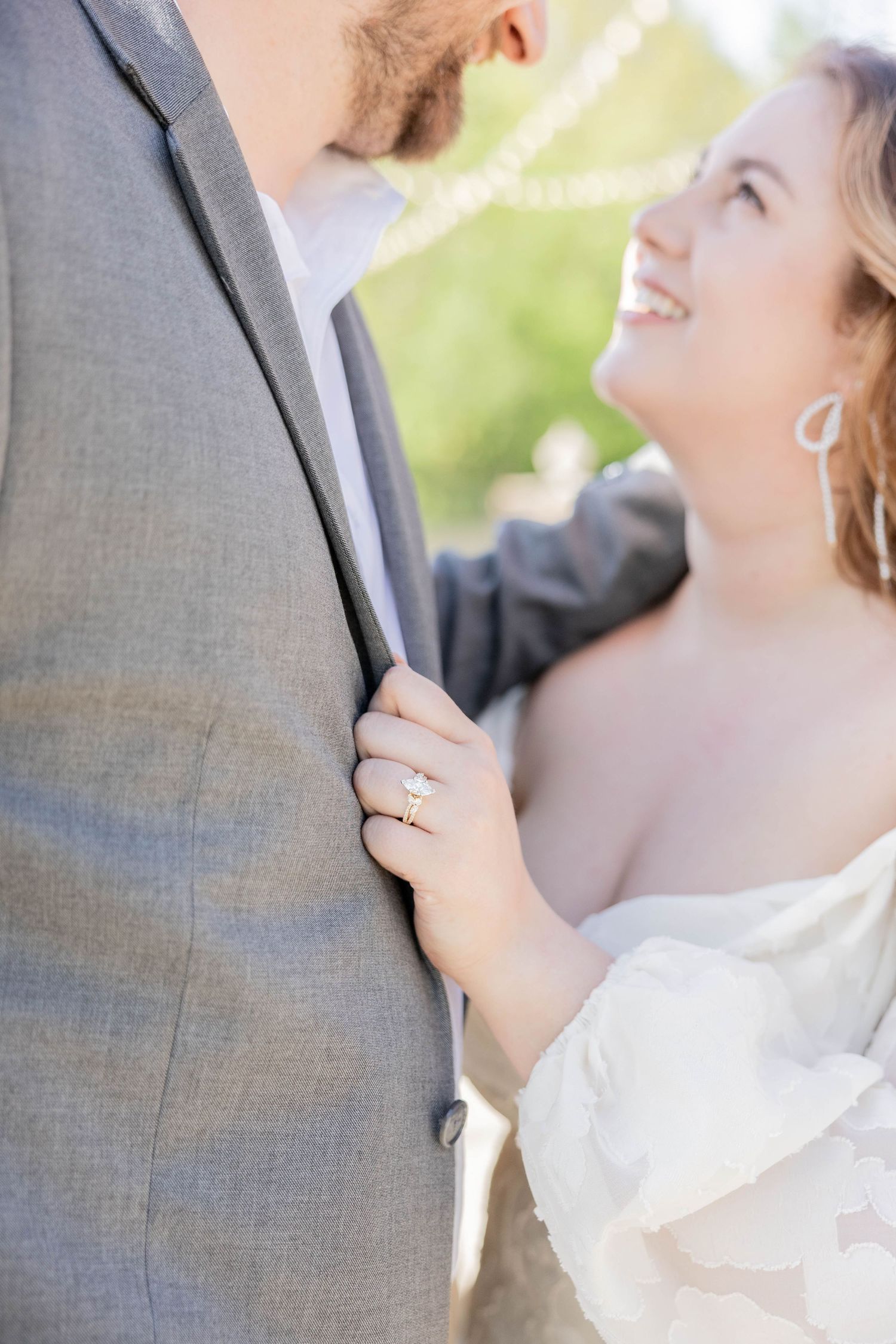 Bride smiling up at groom while holding his gray suit lapel, showcasing her diamond engagement ring outdoors.