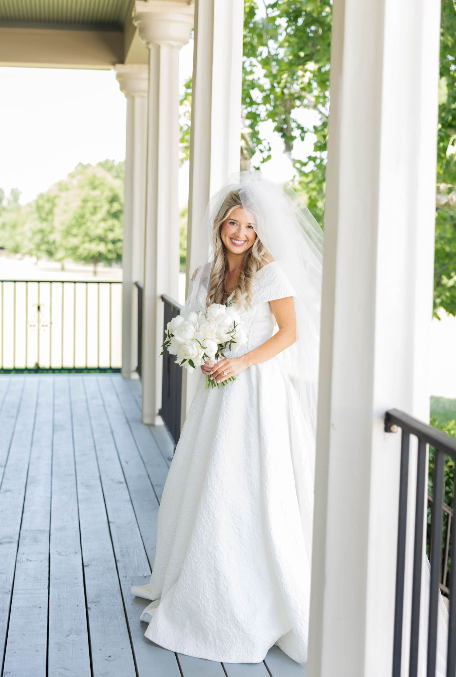 Smiling bride in white lace gown and veil holding white bouquet on a wooden porch with columns.