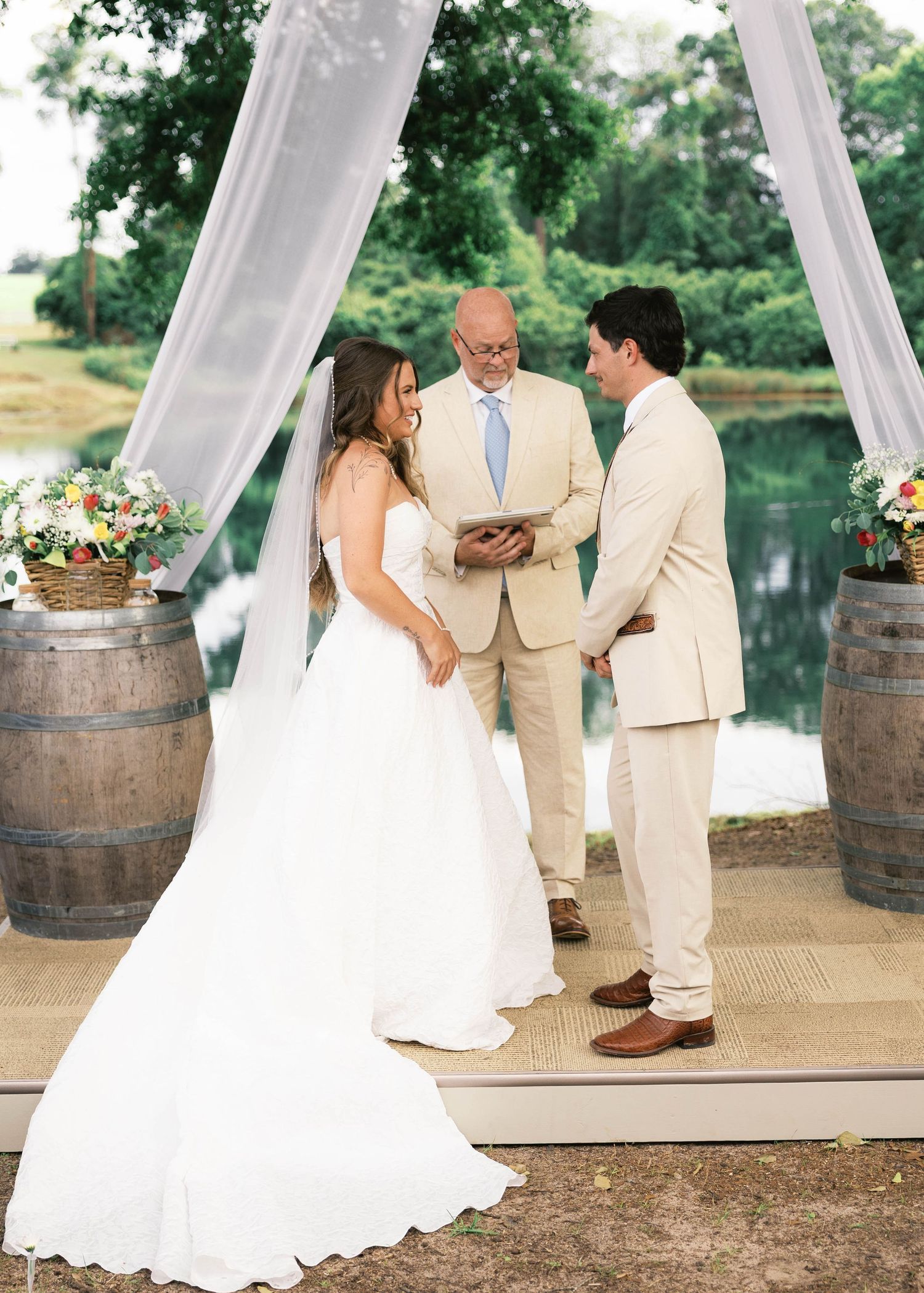 Outdoor wedding ceremony with bride in white gown and groom in tan suit exchanging vows before officiant near rustic barrels.