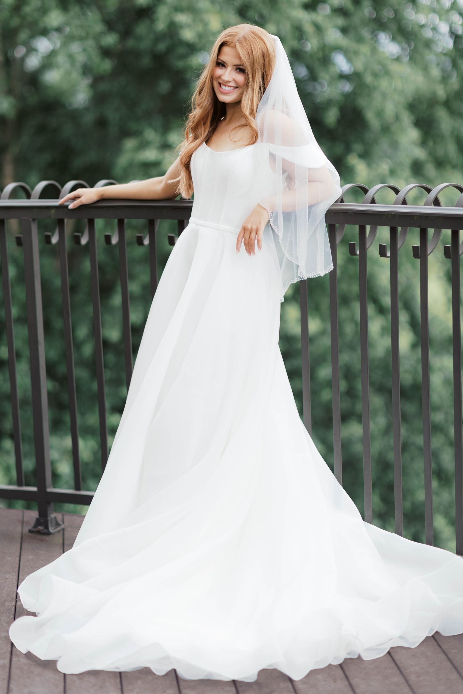 Smiling bride in elegant white A-line gown with veil stands on wooden balcony surrounded by lush green trees.