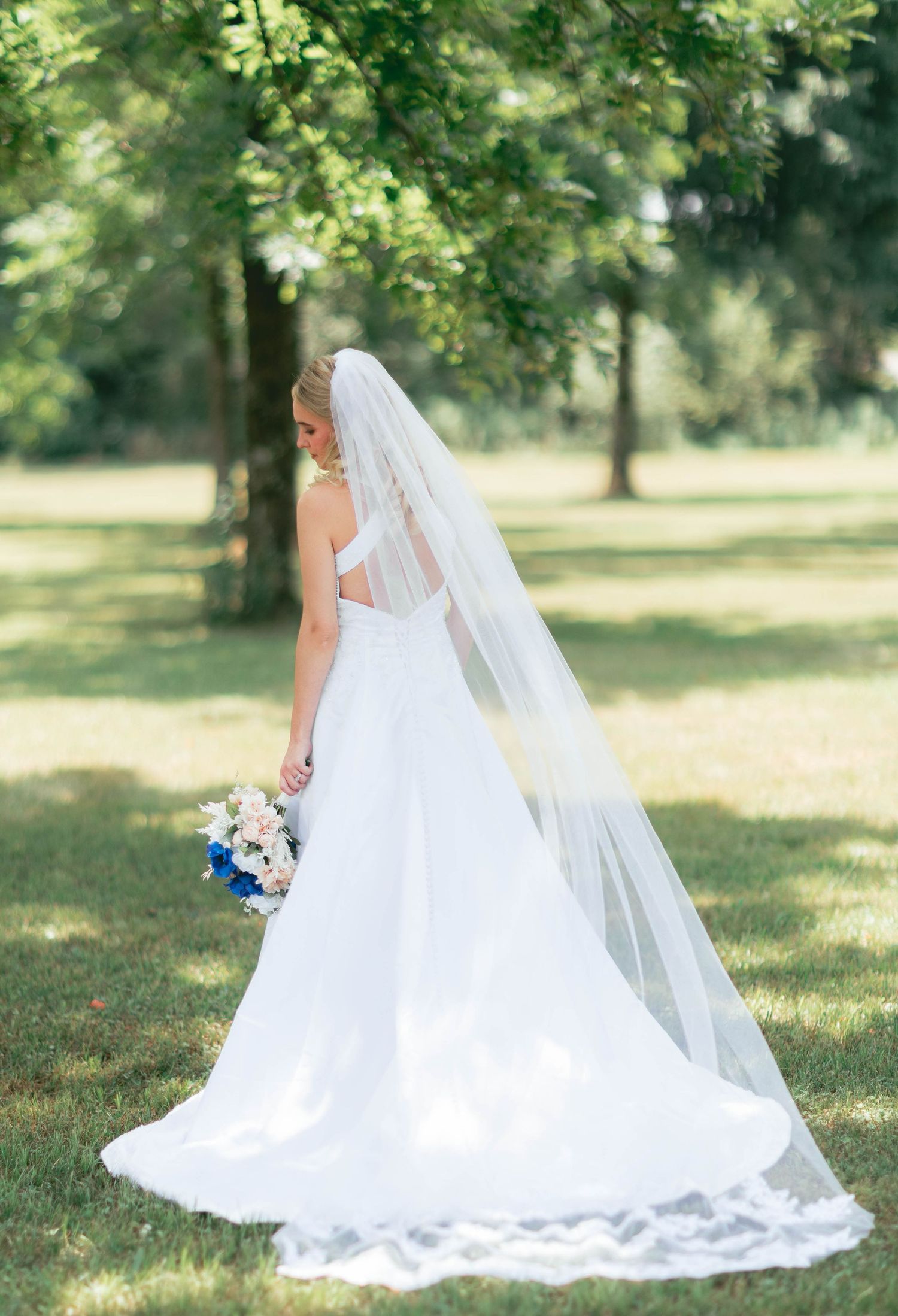 Bride in a stunning white ball gown with lace-trimmed veil holds a bouquet while posing outdoors among green trees.