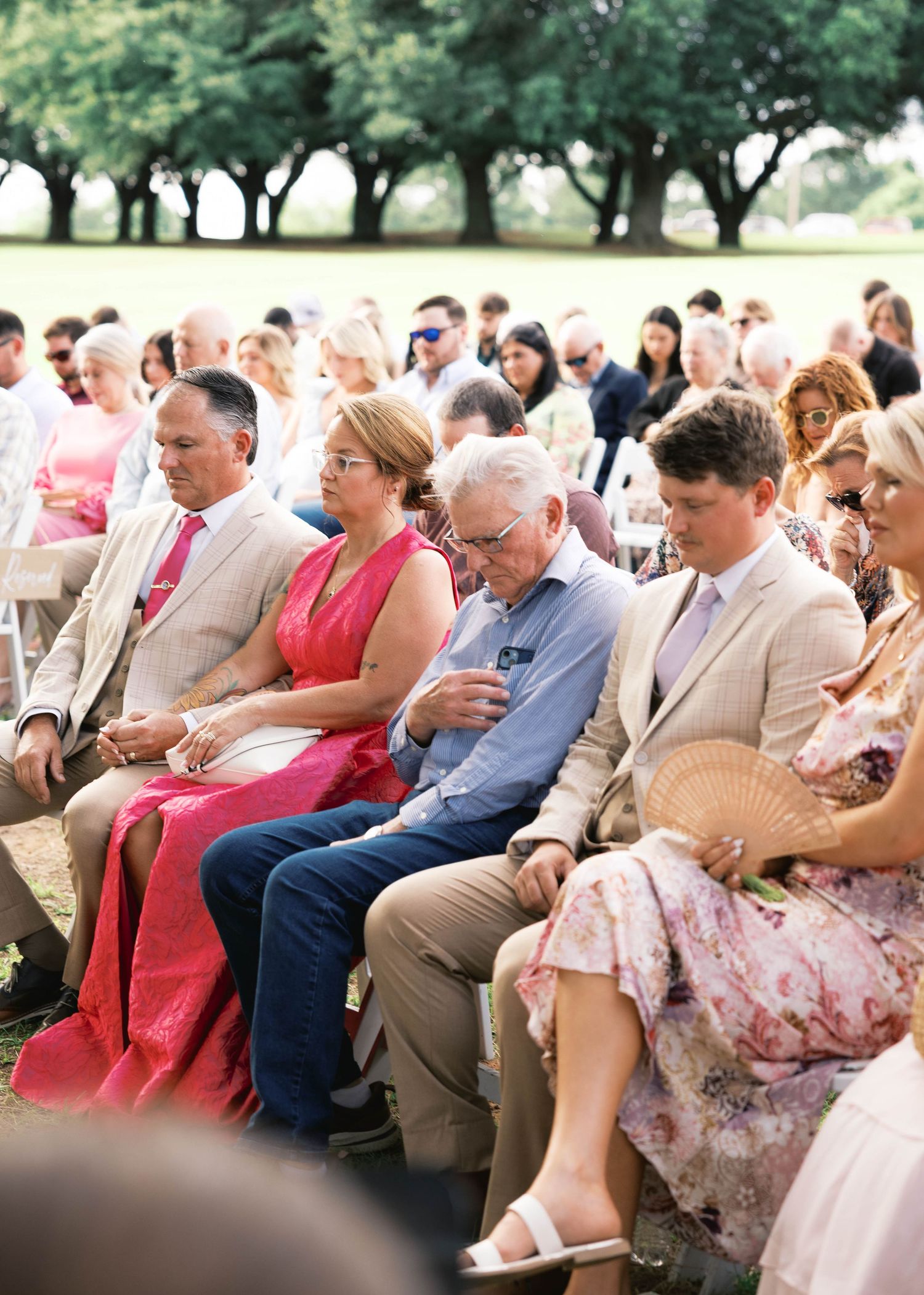 Wedding guests seated outdoors in white chairs during a sunny ceremony, dressed in elegant attire including pink and floral dresses.