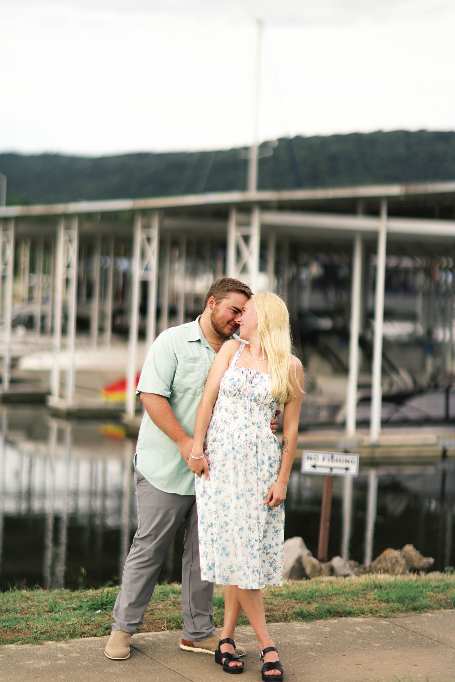 Couple sharing a romantic moment by a marina dock, woman in floral dress and man in mint shirt holding hands.