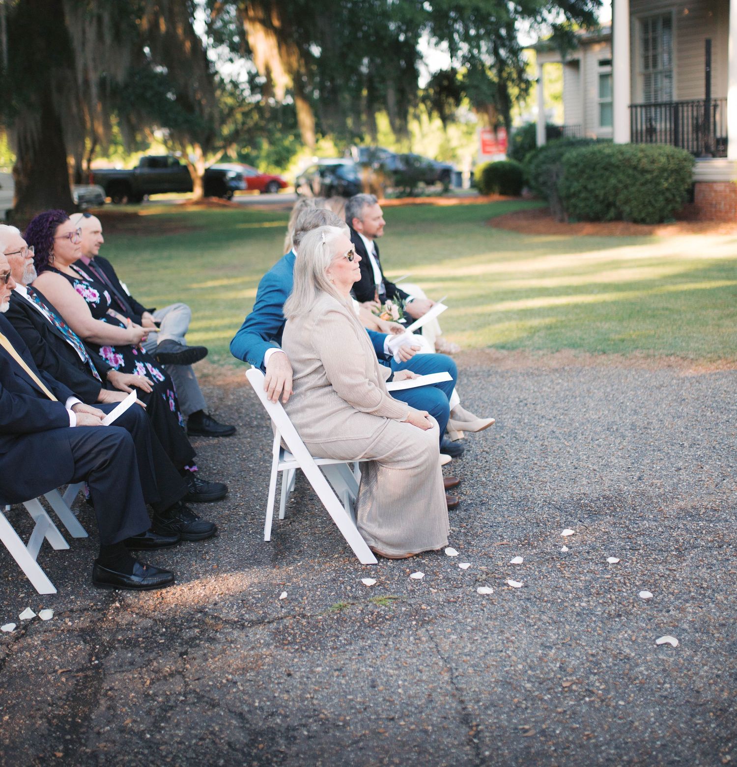 Wedding guests seated in white folding chairs outdoors under large oak trees during a garden ceremony.