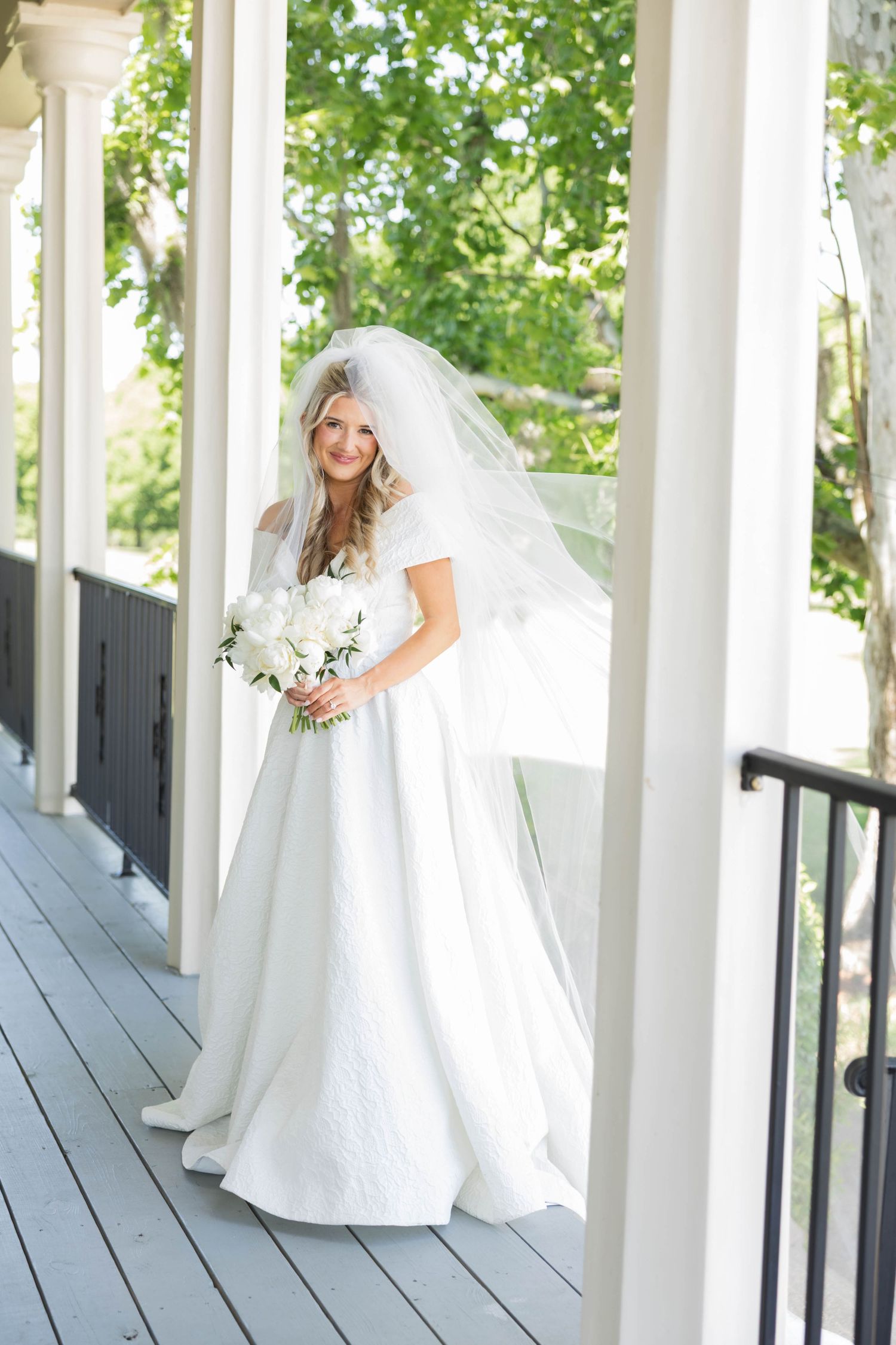 Smiling bride in white gown and veil holding white bouquet on outdoor porch with columns.