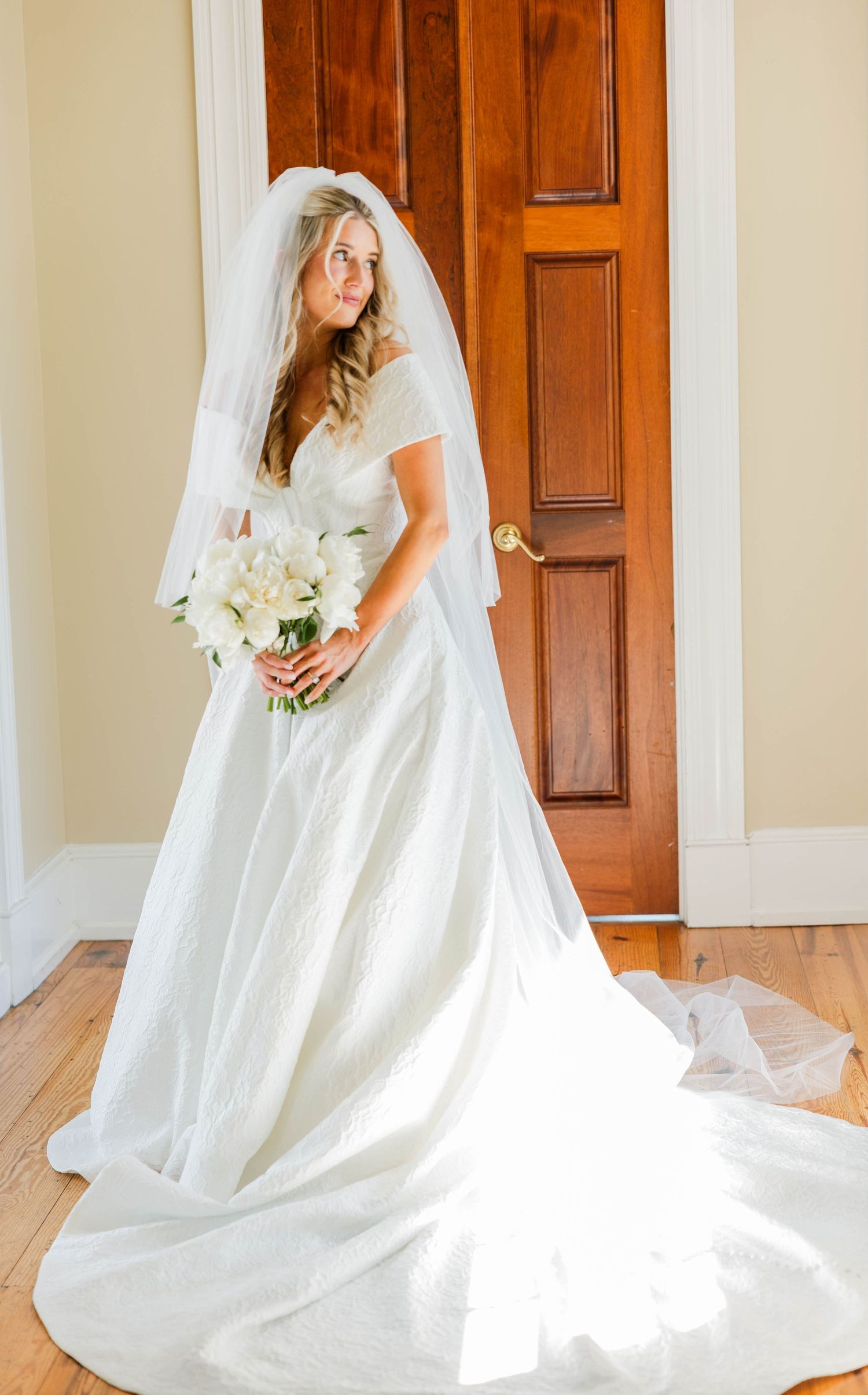 Bride in white wedding gown with veil and bouquet standing near wooden door on hardwood floor.