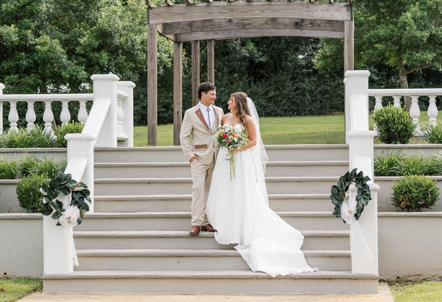 Bride and groom stand on elegant white stone steps under a pergola, surrounded by lush greenery at an outdoor wedding.