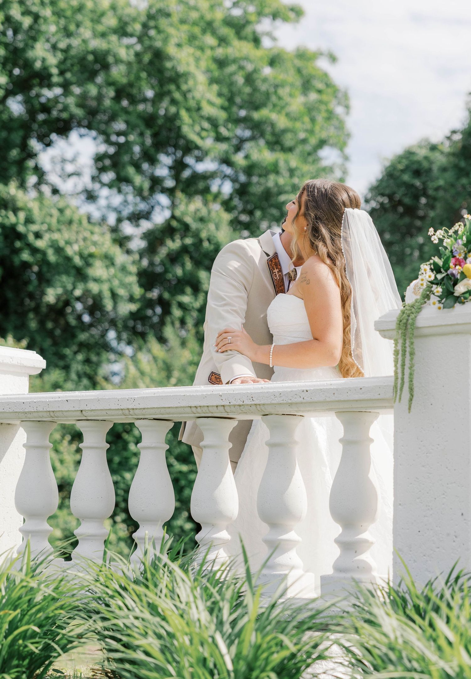 Bride and groom share a romantic kiss on a white balustrade balcony surrounded by lush green trees.