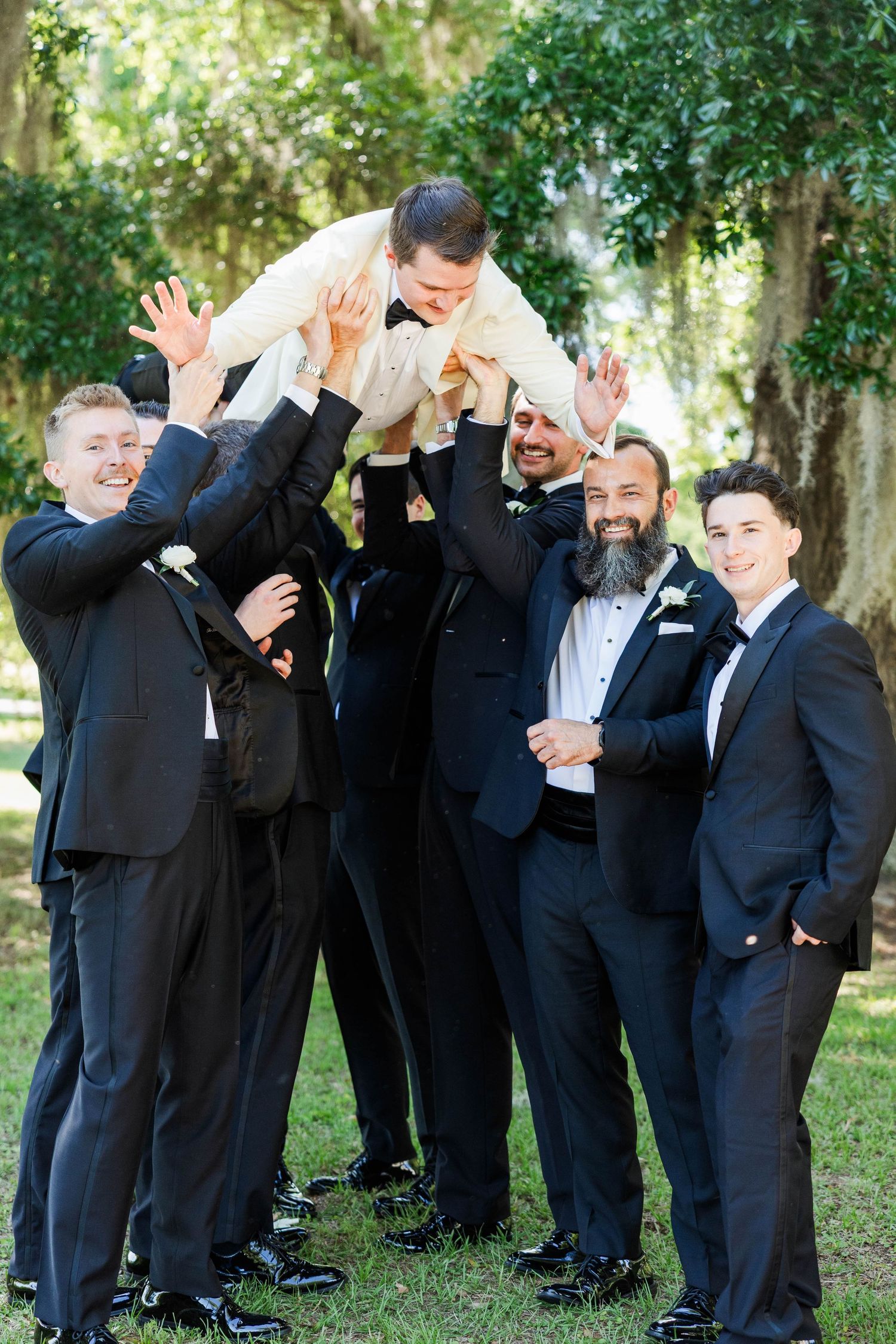 Groomsmen in black tuxedos lifting the groom in a cream jacket outdoors at a wedding celebration.