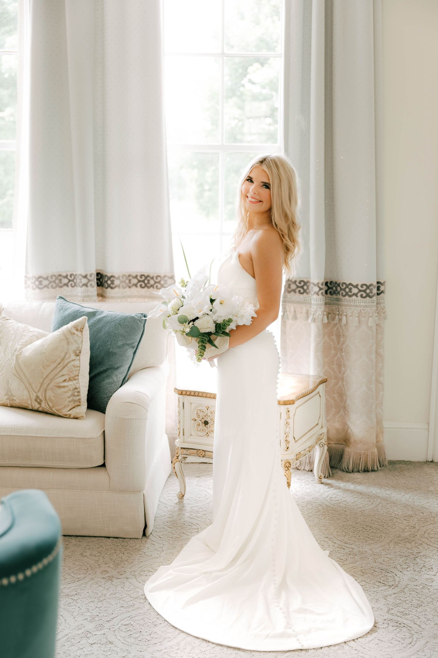 Blonde bride in sleek white gown holds white bouquet by bright window in elegant room.