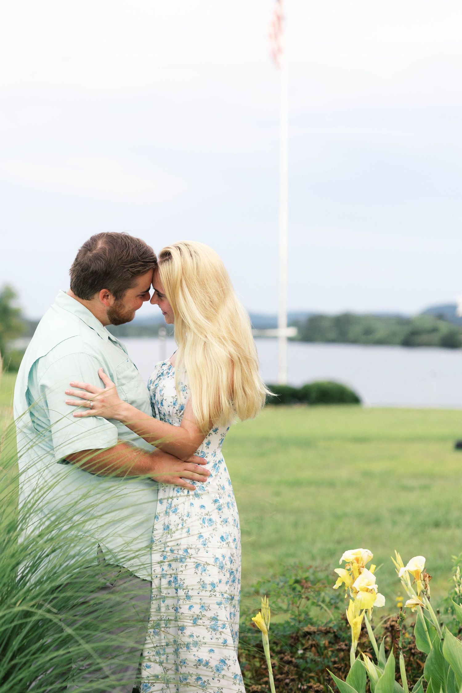 Couple sharing a romantic moment outdoors near a waterfront garden with yellow flowers and a flagpole.