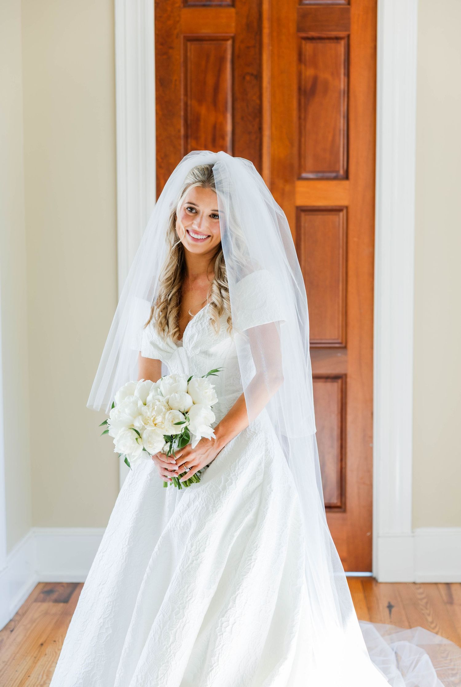 Bride in white wedding gown and veil holding white floral bouquet, smiling in front of wooden doors.