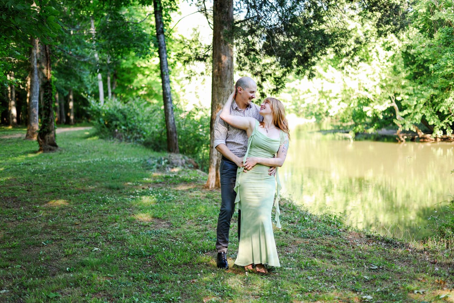 Couple embracing outdoors near a serene pond surrounded by lush green trees on a sunny day.