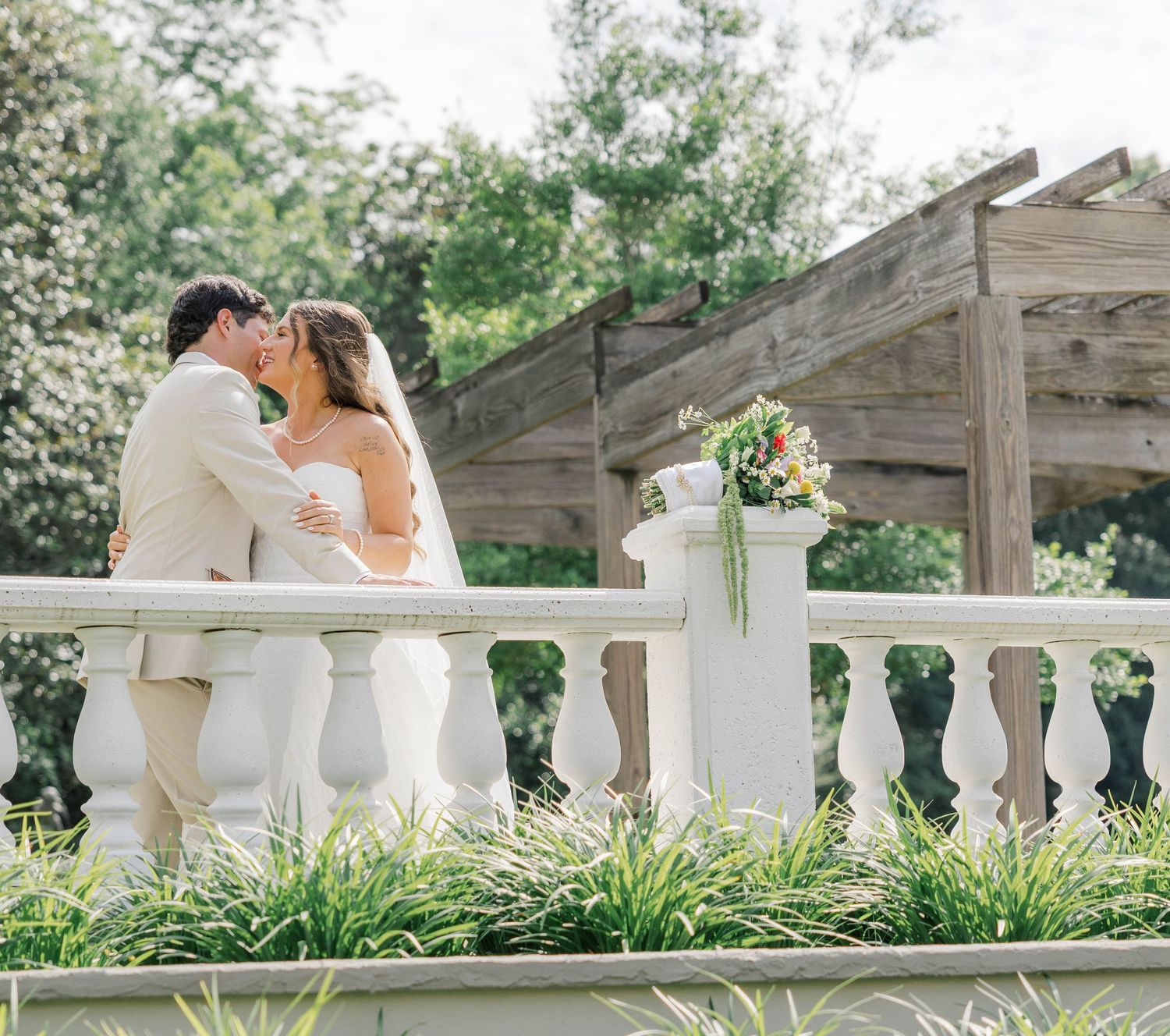 A bride and groom share a romantic kiss on a white balustrade terrace surrounded by lush greenery with a floral bouquet nearby.