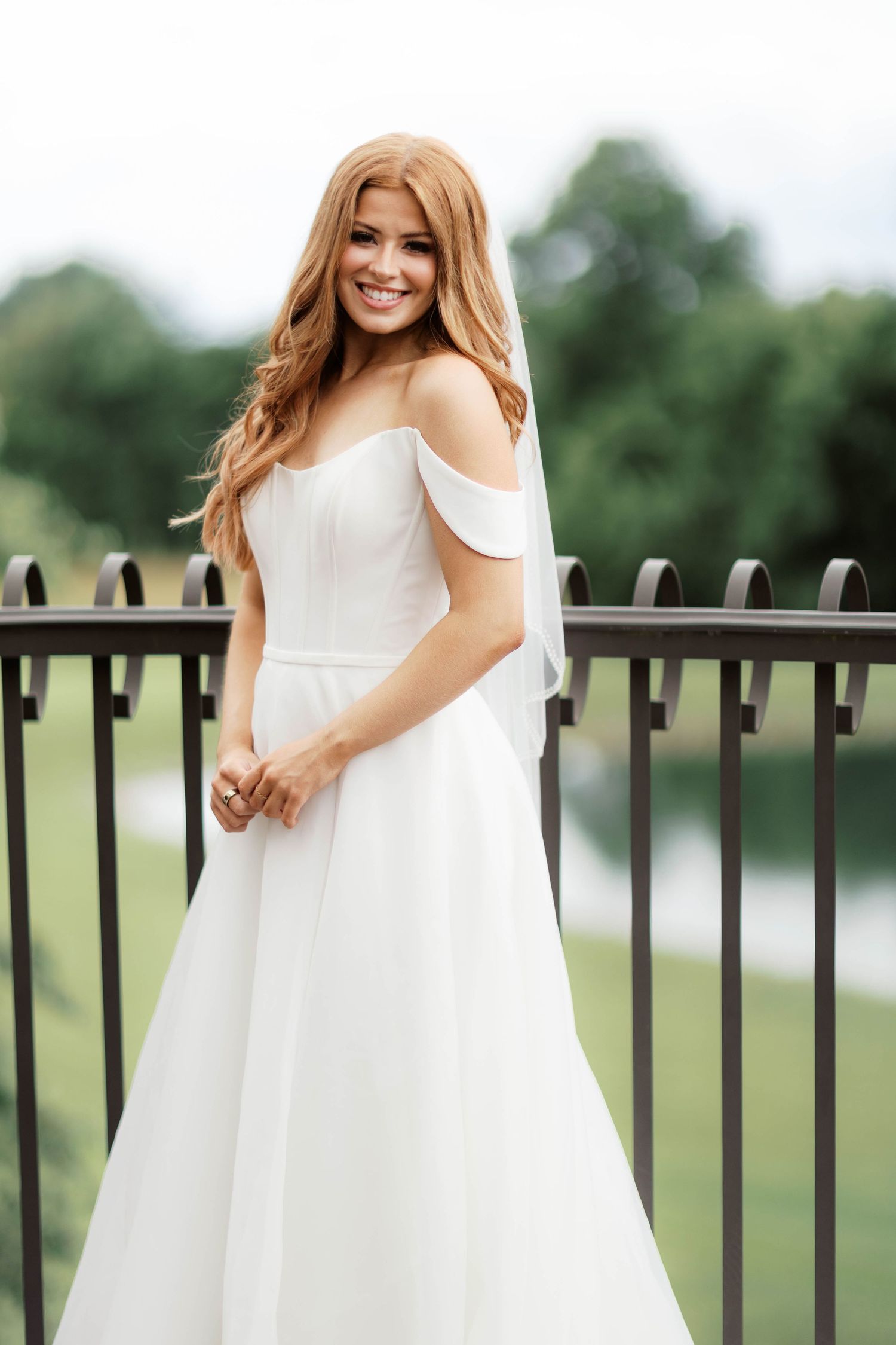 A smiling bride in an off-shoulder white A-line wedding gown stands on a balcony with lush greenery in the background.