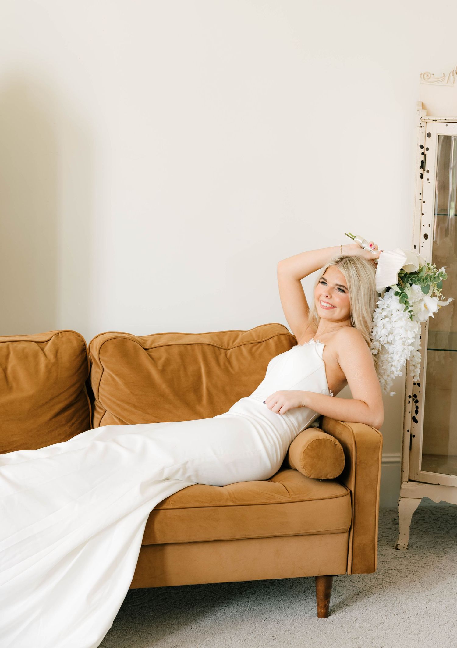 Blonde bride in white gown relaxing on a caramel velvet sofa with floral decor nearby.